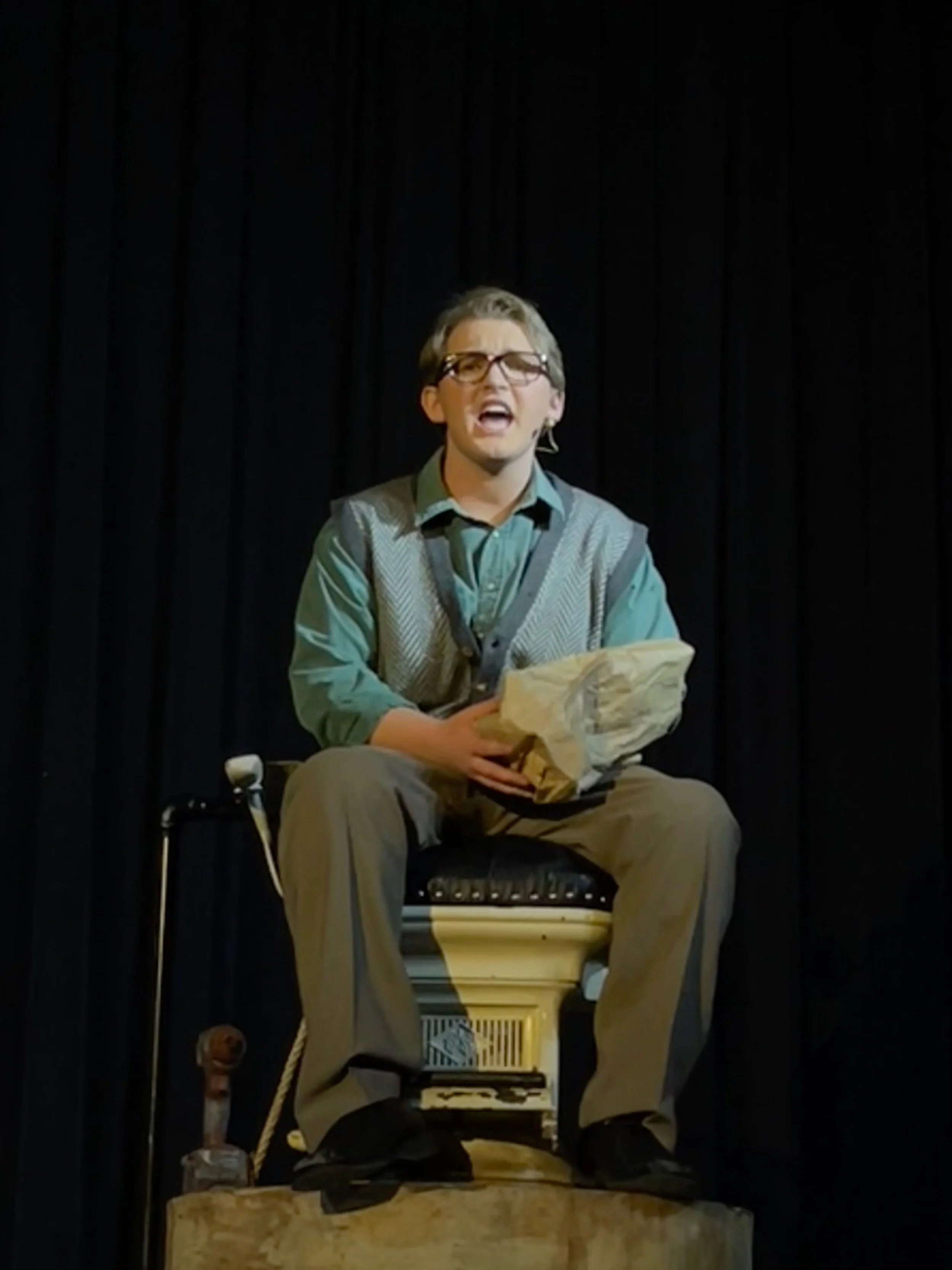 Man performing on stage, sitting on a toilet, holding a bag, wearing glasses, with a black curtain backdrop.