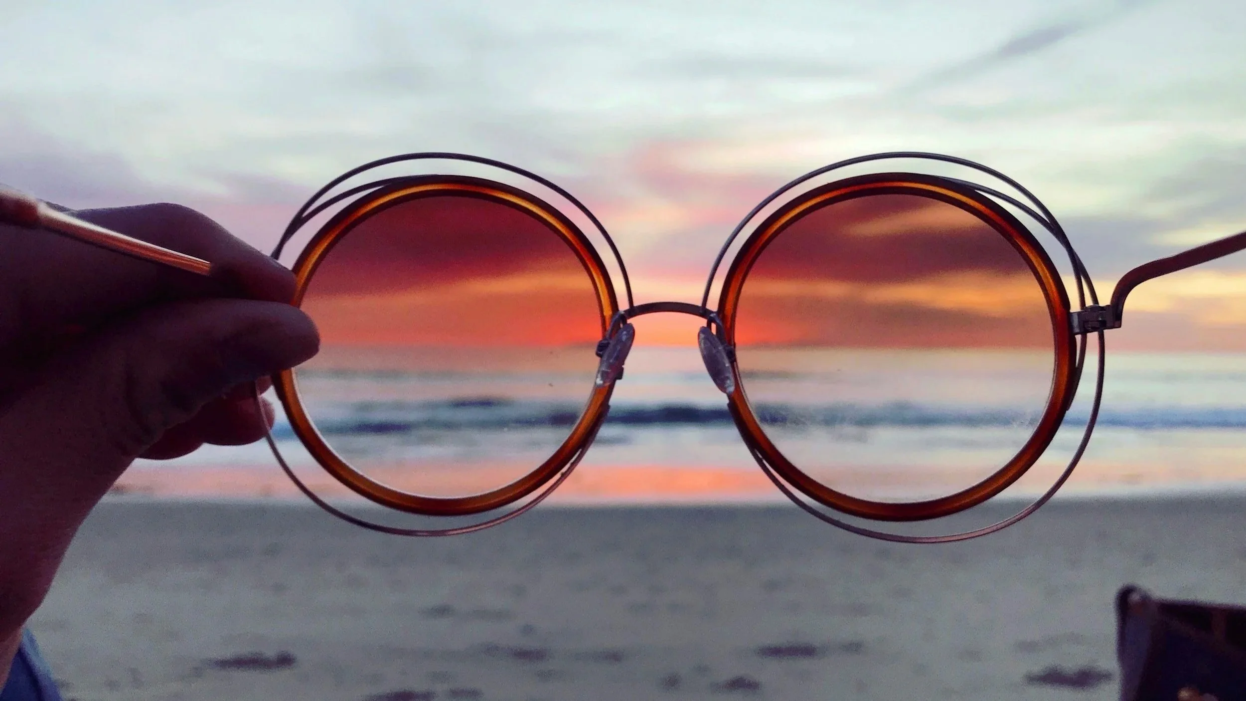 Tortoise shell sunglasses held up against sunset at the beach with waves and colorful sky.