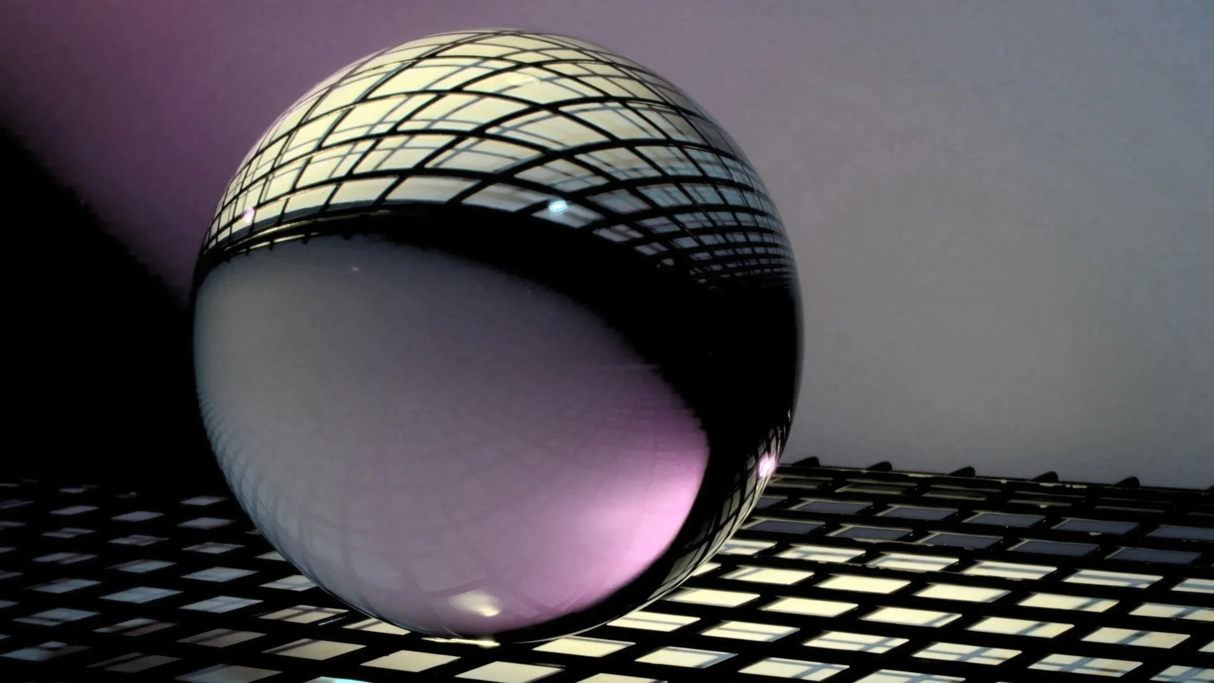 A highly reflective silver sphere resting on a grid-patterned surface, reflecting a geometric ceiling structure and ambient light.
