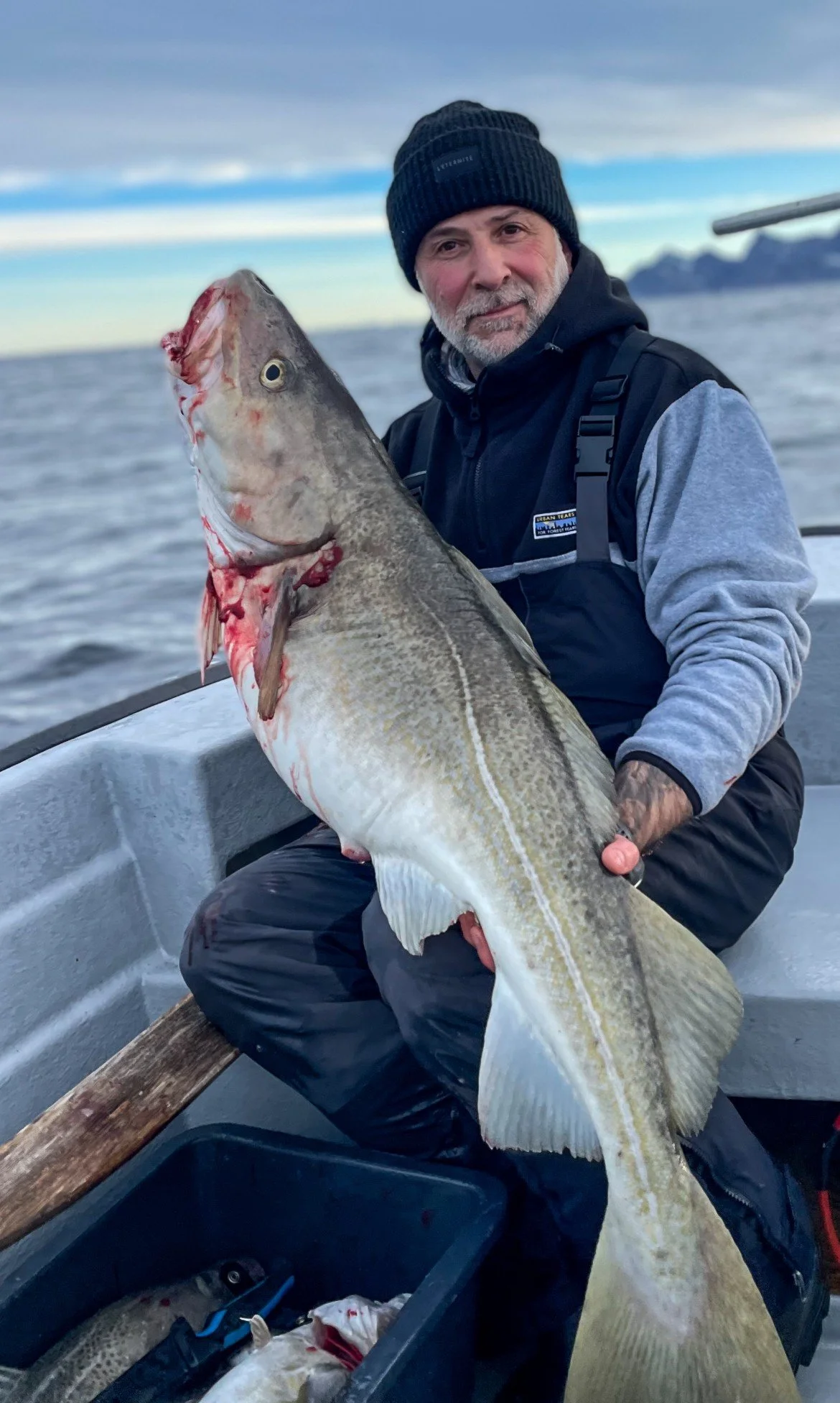 En mann med grått skjegg og svart lue sitter i en båt og holder et storfisk med begge hender. Det er vann og fjell i bakgrunnen.