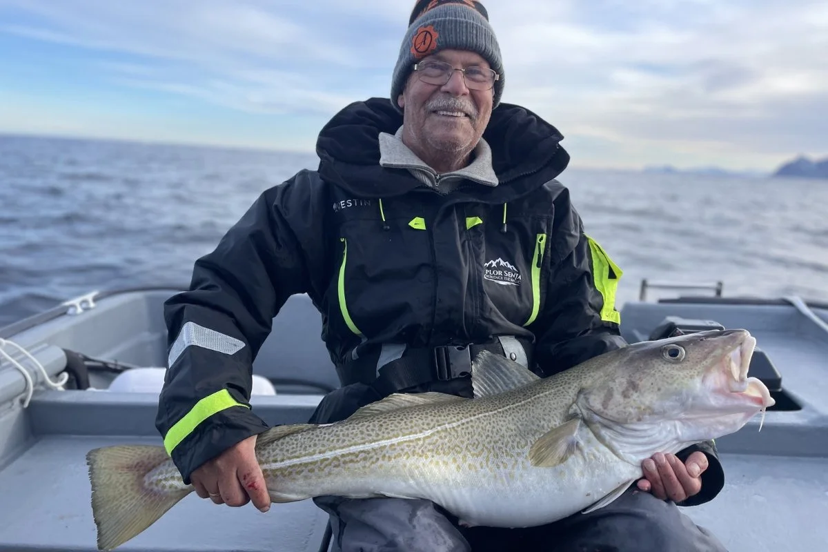 Person som holder opp en stor fisk på en båt til sjøen, iført ullhue, jakke og hansker, gir tommel opp.