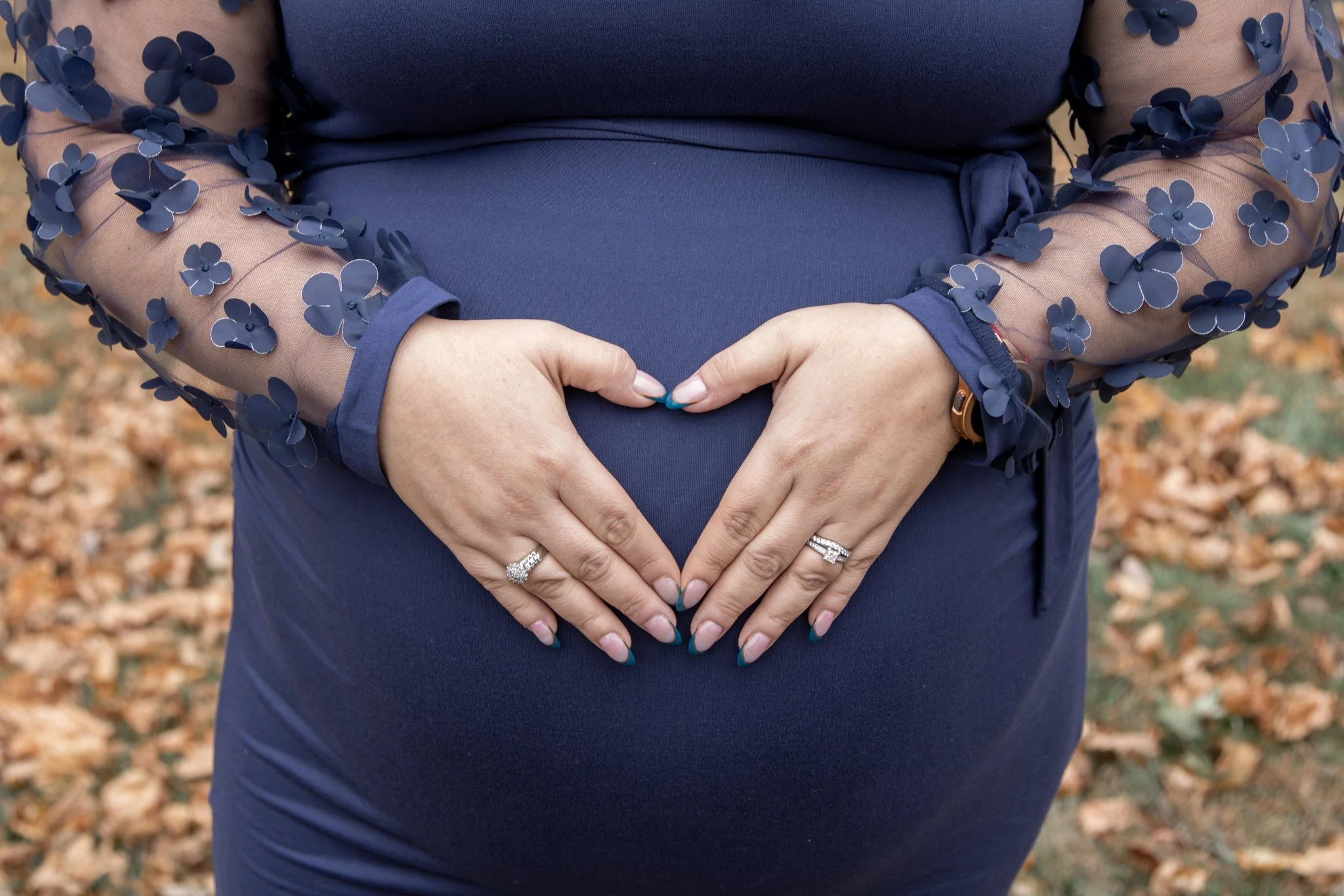 A person in a navy blue dress with sheer sleeves decorated with 3D floral appliqués forms a heart shape with their hands over a pregnant belly. The person wears rings and a bracelet, and the background shows fallen autumn leaves.