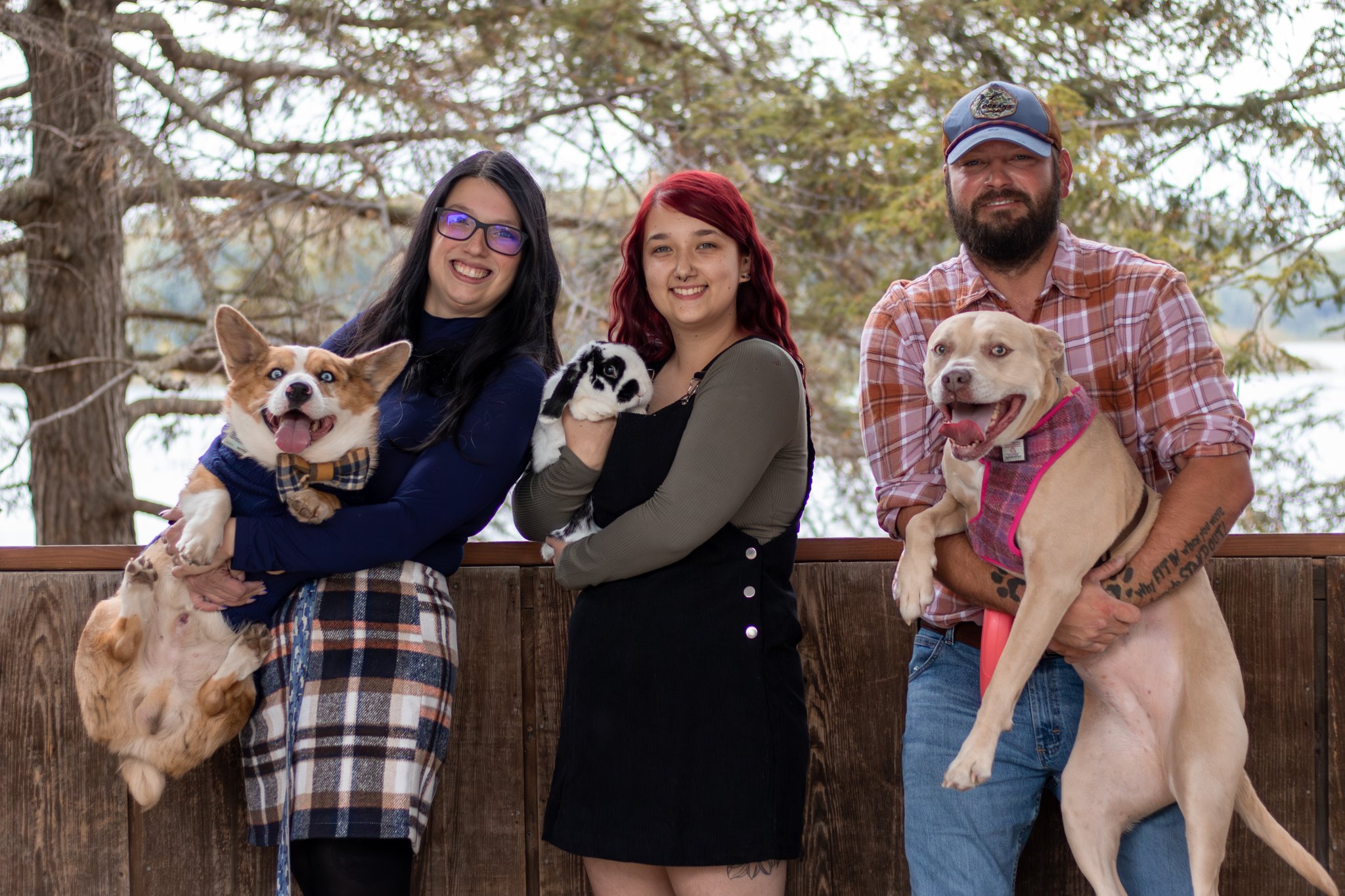 Three people standing outdoors with trees in the background, each holding a dog and a plush rabbit toy.