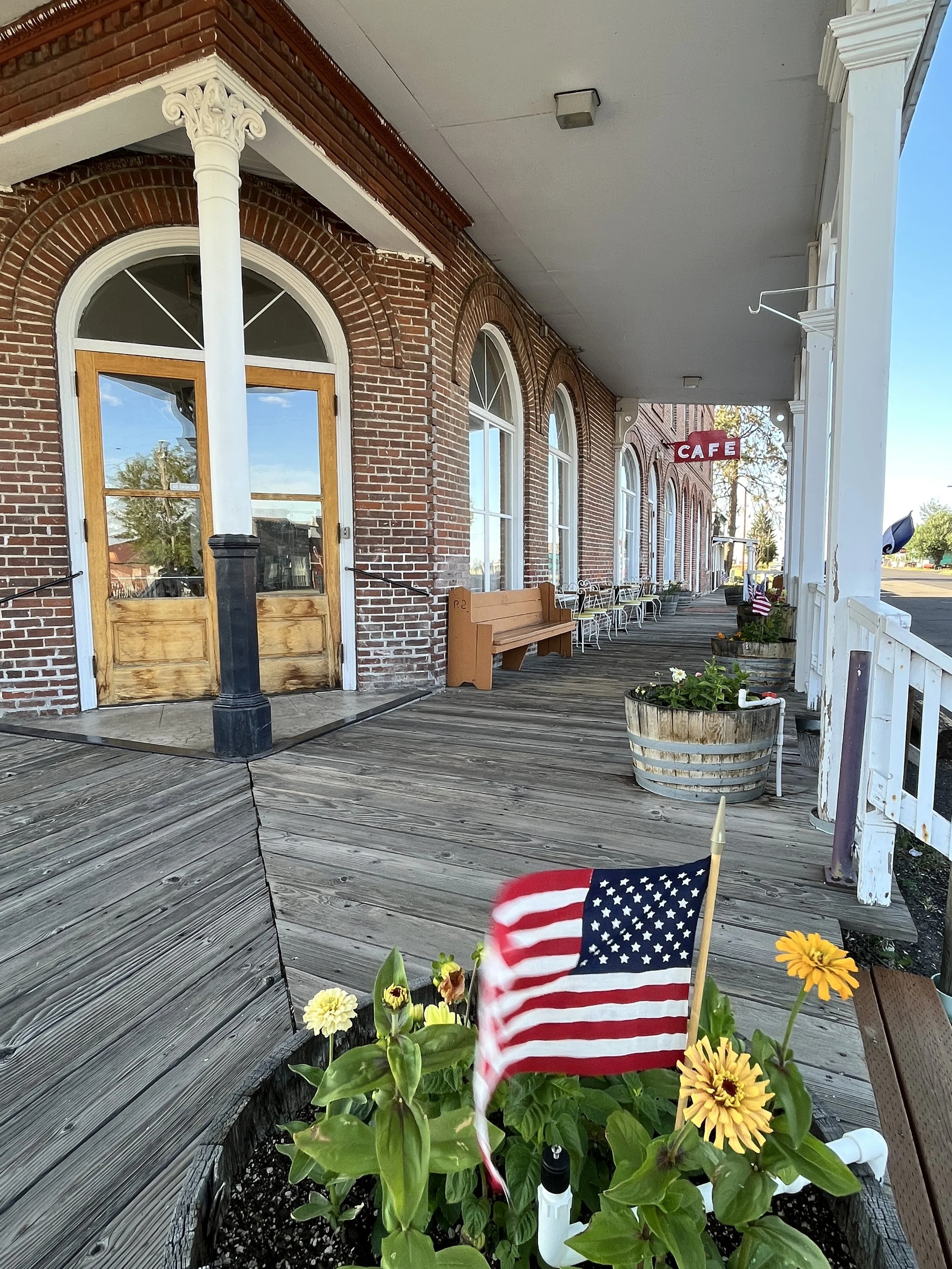 Front porch of a historic brick building with large arched windows and a café sign. Flower pots and outdoor seating are visible, along with a small plant adorned with an American flag in the foreground.