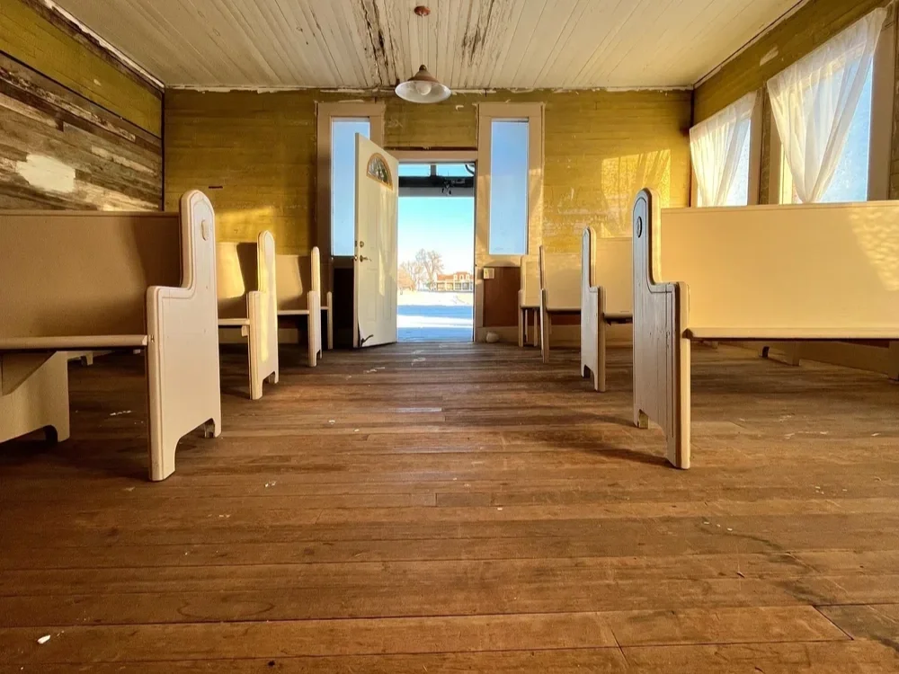 Empty room with wooden floors, cream-colored benches along the sides, open door showing a winter scene outside, sunlight streaming through windows with white curtains.