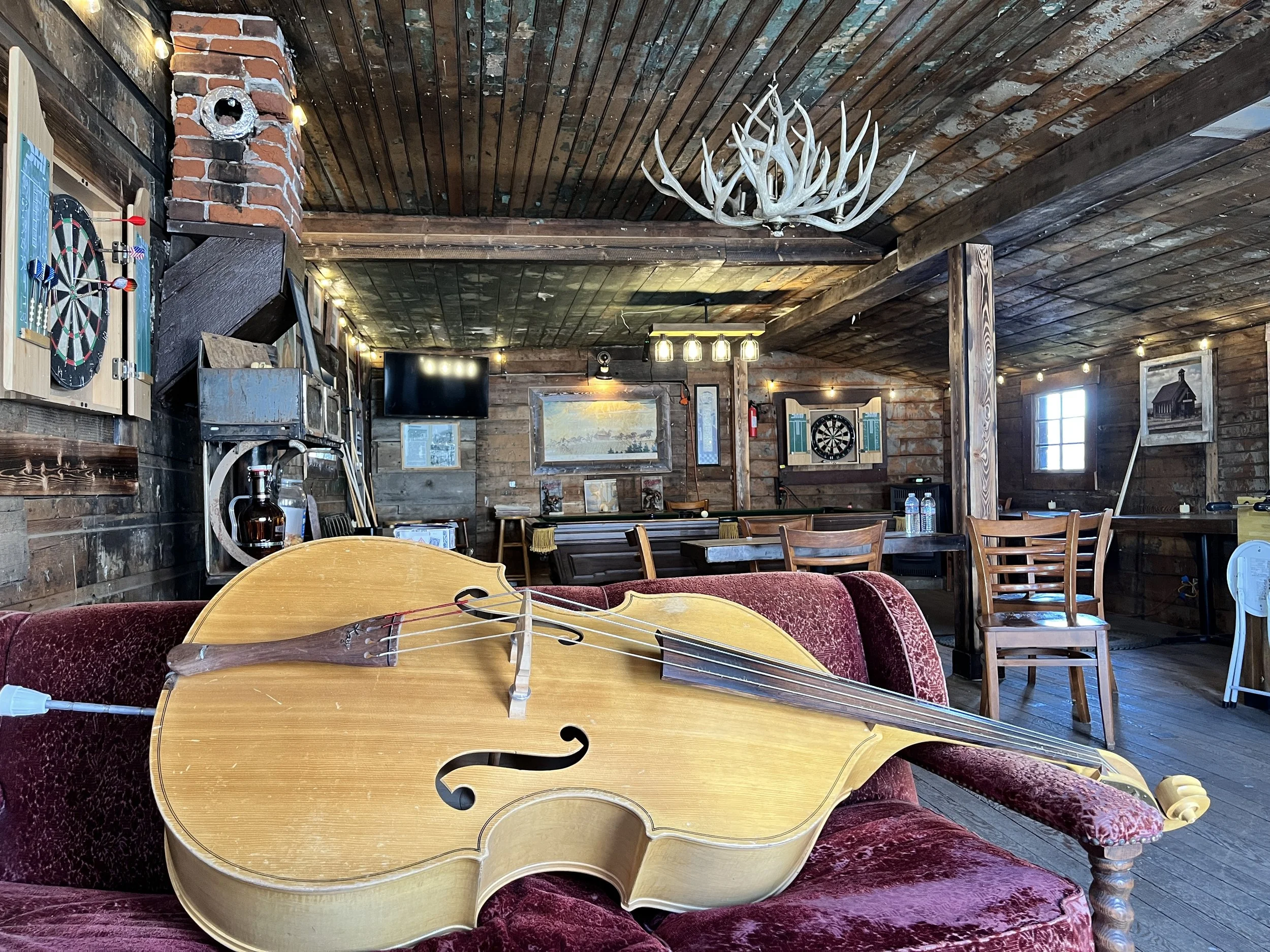 An interior of a rustic wooden room with a large acoustic guitar on a velvet sofa, dartboards on the wall, a chandelier made of antlers, tables and chairs, and various decorations.