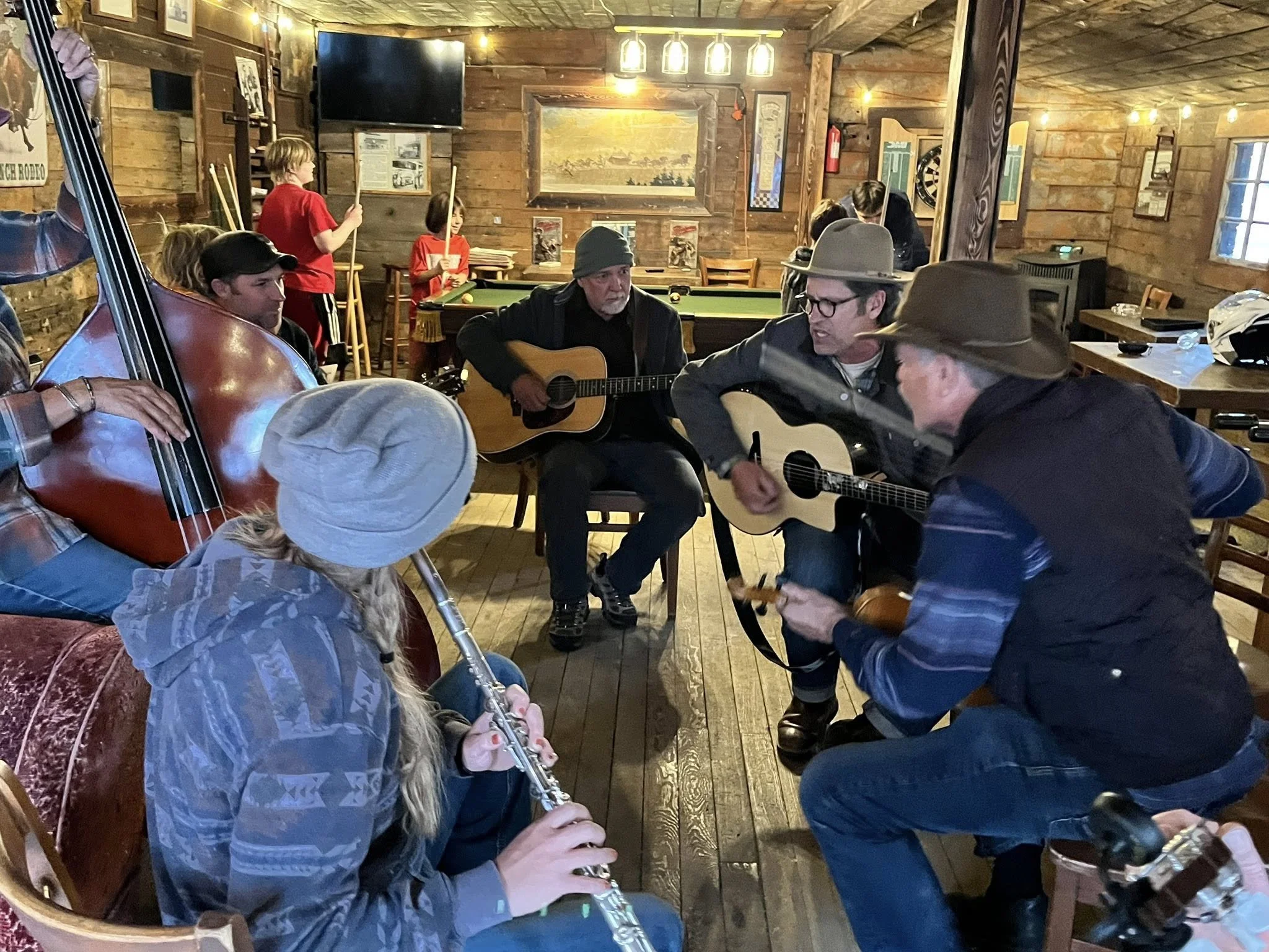 Group of musicians playing guitars and a flute in a cozy, rustic wooden room with children in the background, some near a pool table and others reading or standing.