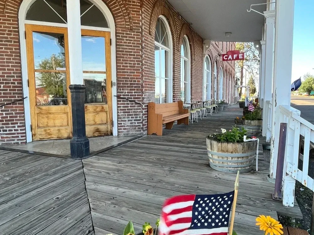 Small outdoor dining area with wooden deck, brick building with large arched windows, wooden door, benches, tables, and potted plants. An American flag is in the foreground, and a red sign with white letters spelling 'CAFE' hangs on the building.