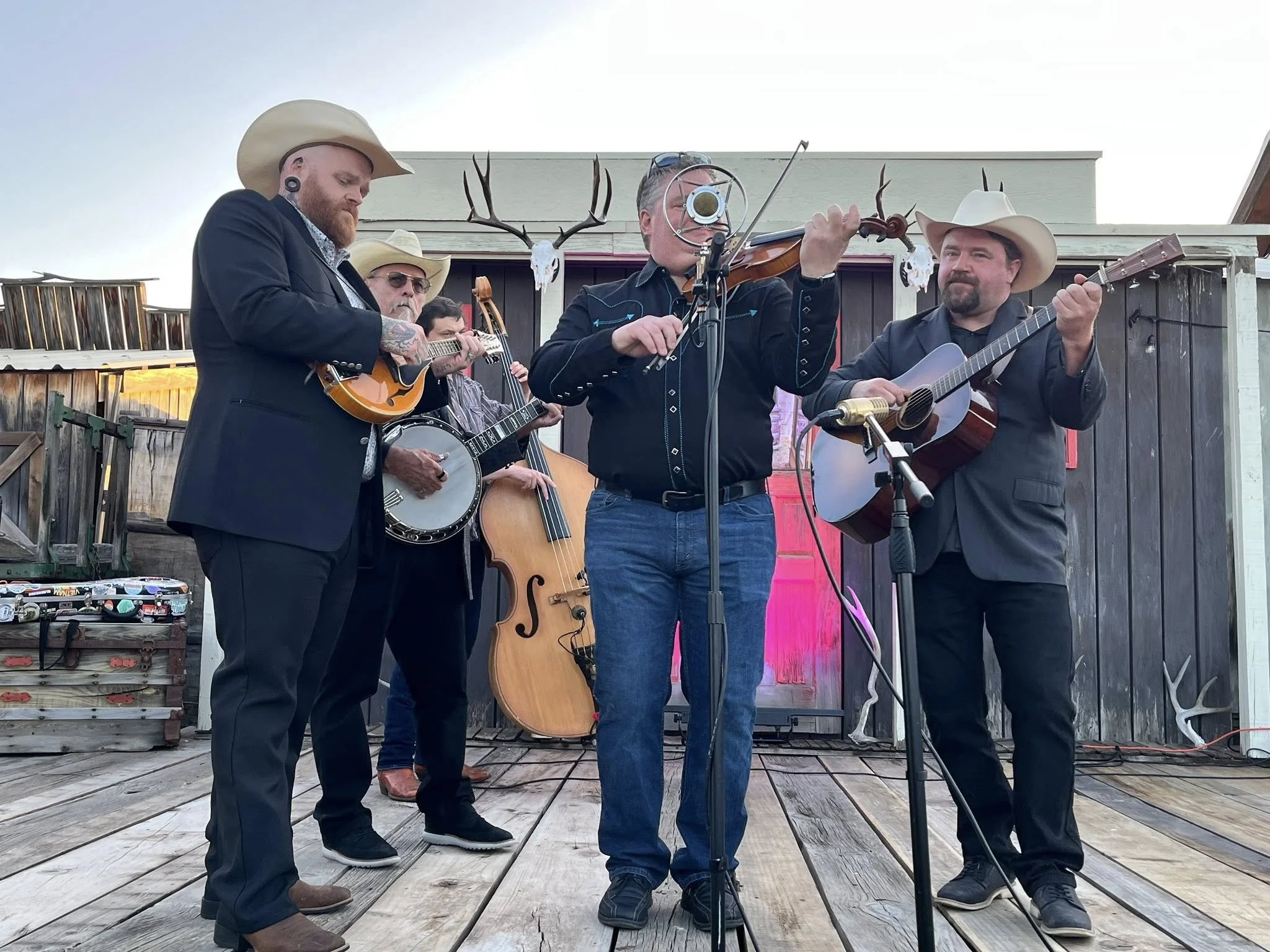A group of five men performing music on an outdoor stage, all wearing cowboy hats and dark suits, with one man playing a violin, another a mandolin, and others playing guitars, with a rustic wooden backdrop and antlers mounted on the wall.