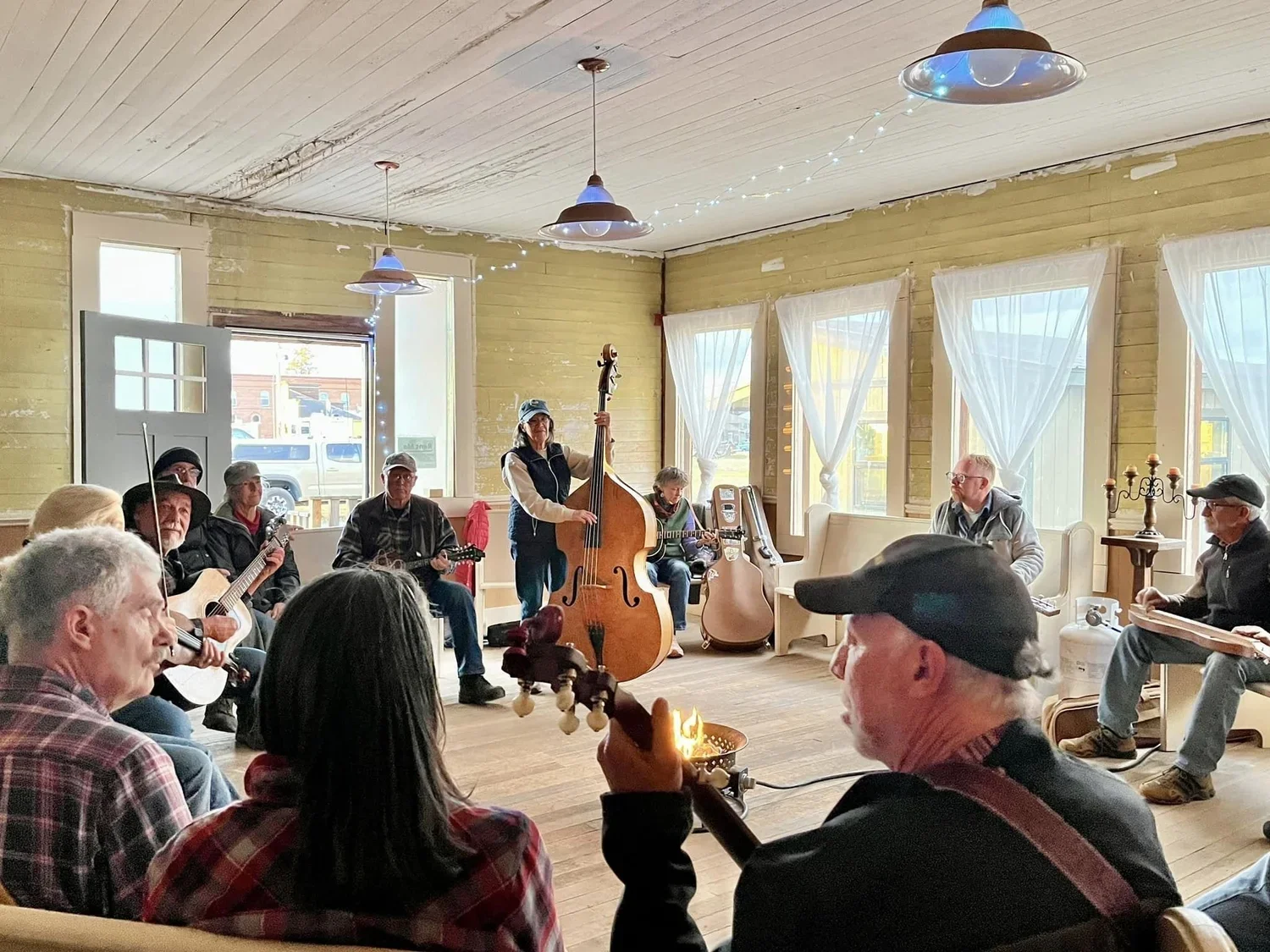 A group of musicians performing in a cozy room with large windows and white curtains, some playing guitars, a double bass, and an accordion, while an audience listens.