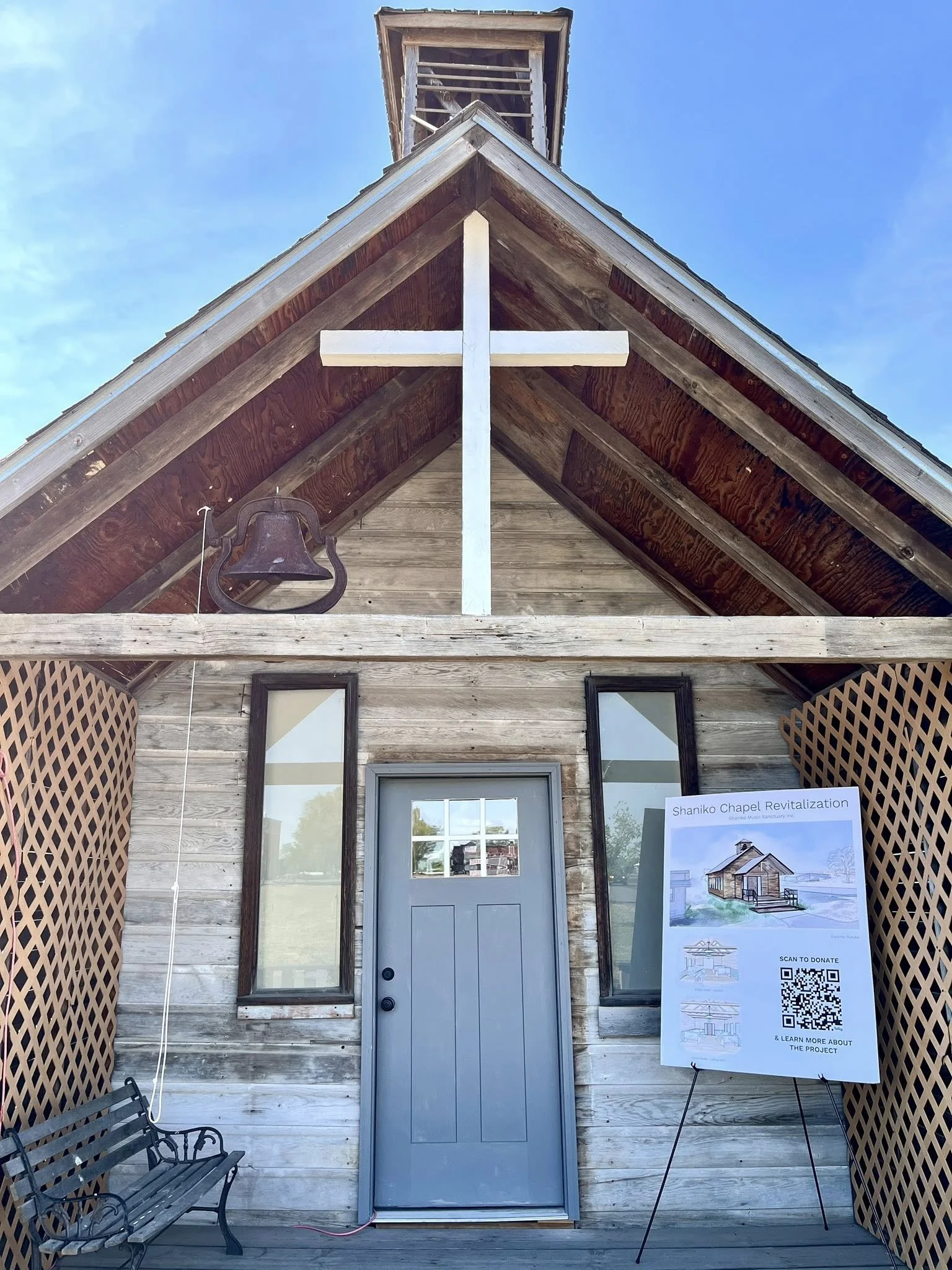Front view of a rustic wooden chapel with a cross mounted above the door, a bell hanging to the left, and a sign on an easel reading "Shaniko Chapel Revitalization" with a QR code.