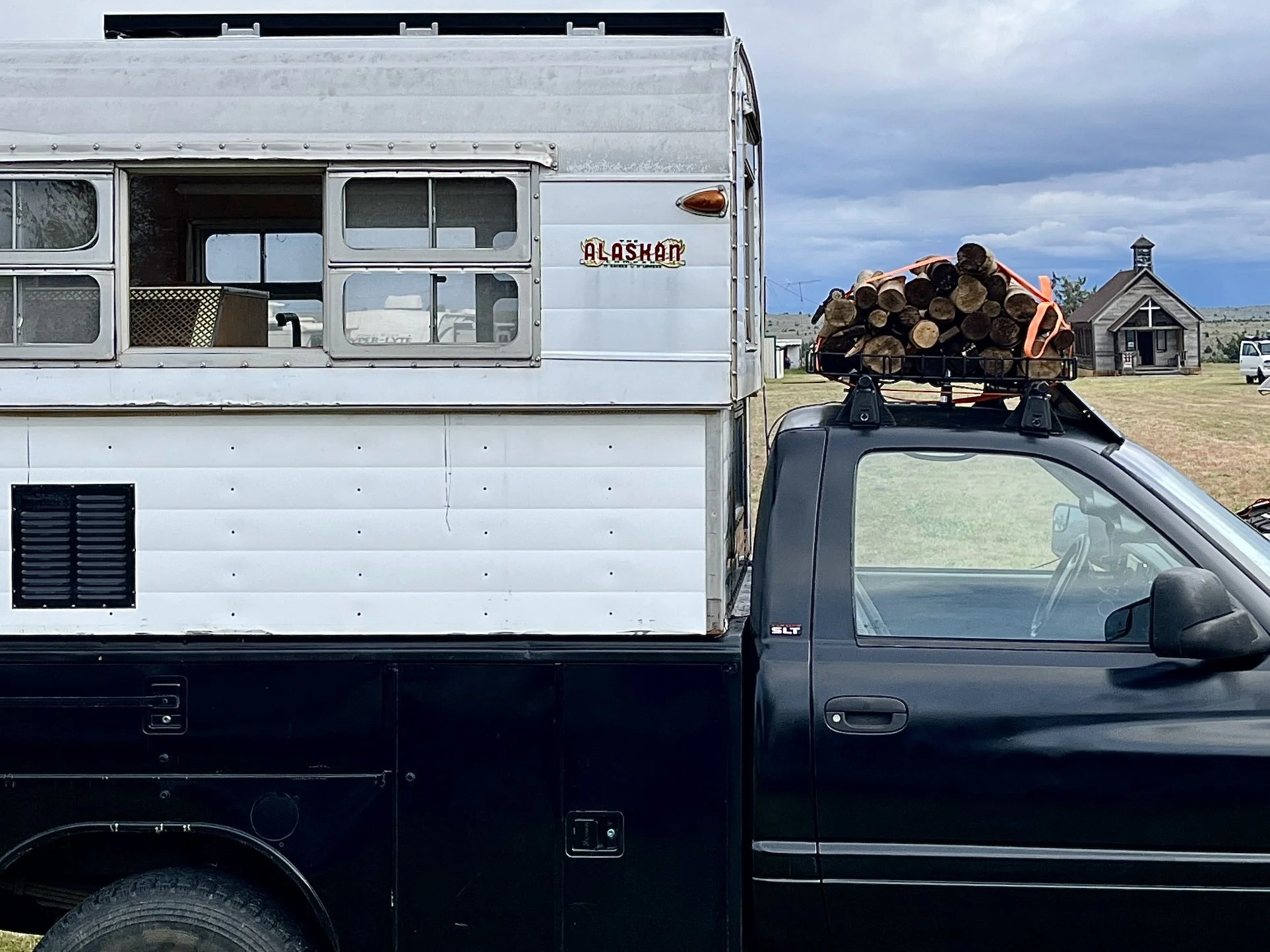 A black pickup truck with a large camper attached, carrying a load of logs on its roof, parked on a rural field with a church in the background.