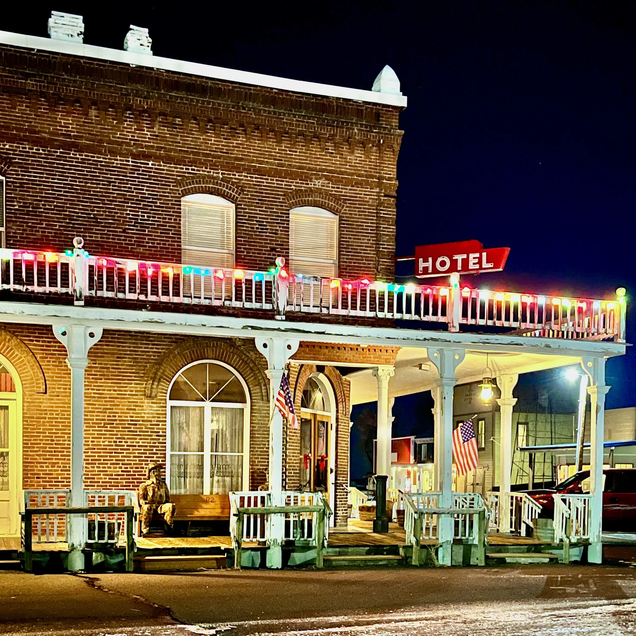 Night view of a brick hotel with a porch, American flags, and colorful string lights. A 