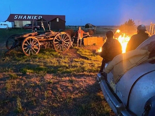 A group of people standing near a campfire at dusk, with an old wagon and a building labeled 'SHANIKO' in the background.