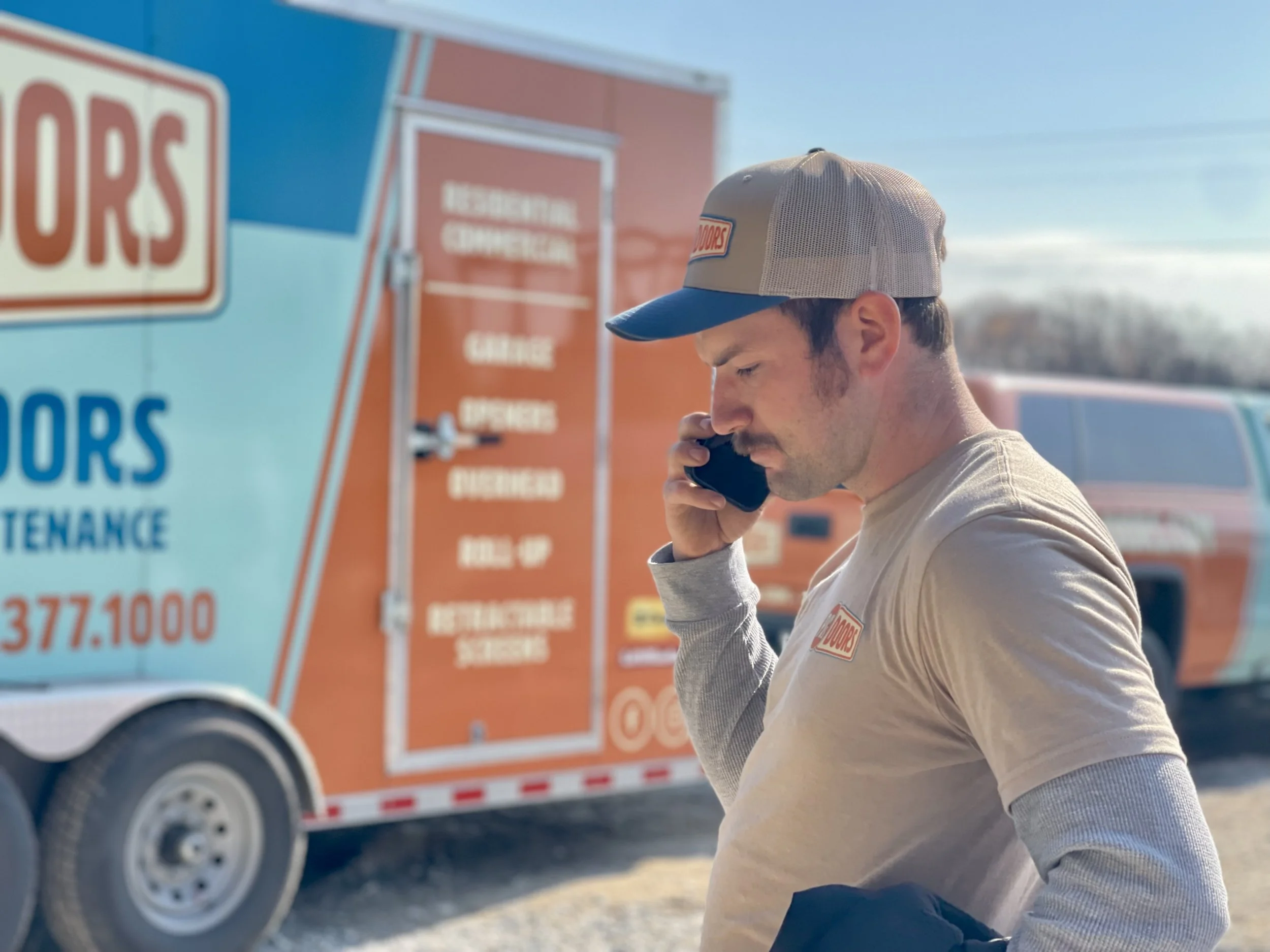 Two smiling men taking a selfie indoors, with a garage door in the background. One wears a black cap, and the other wears a gray cap with a 'New Horizons' logo.