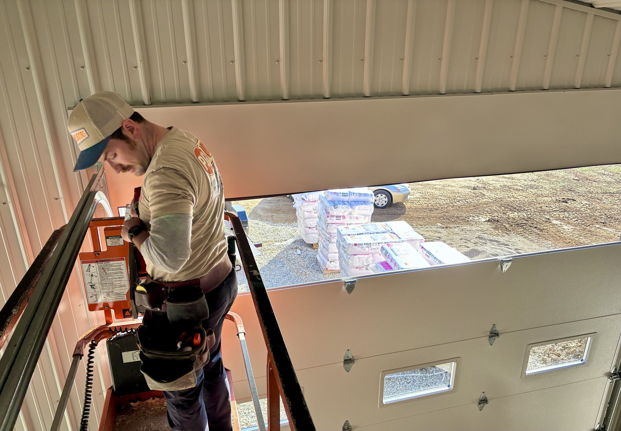 A worker in a beige shirt and cap on an orange lift truck inside a building, with an open garage door revealing stacks of supplies outside.