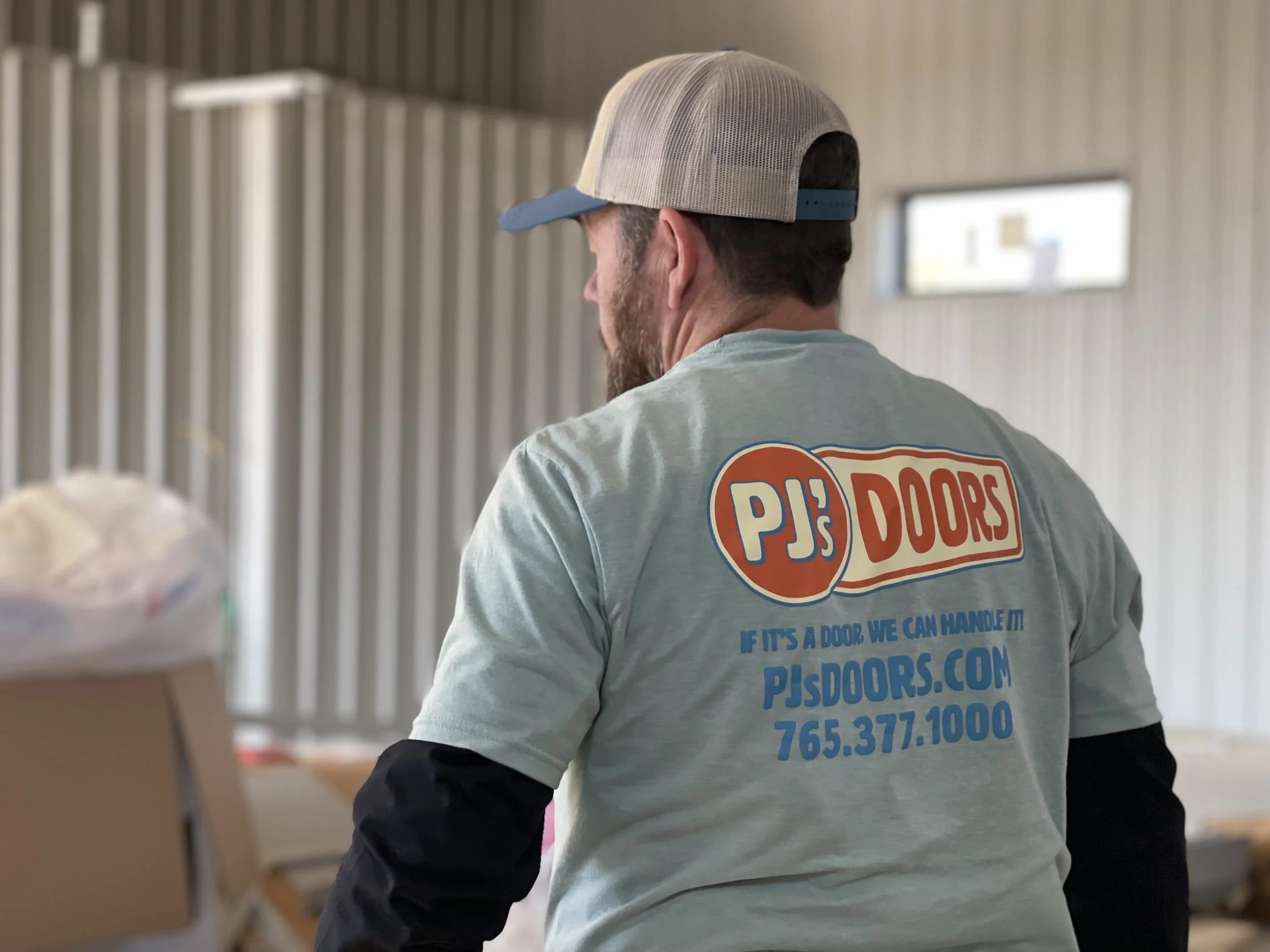 Two workers on a yellow scissor lift installing drywall on an overhead ceiling of a garage. One worker is wearing a black helmet, and the other is wearing glasses and a blue jacket. The garage door is open with a view of a parking lot and neighboring buildings.