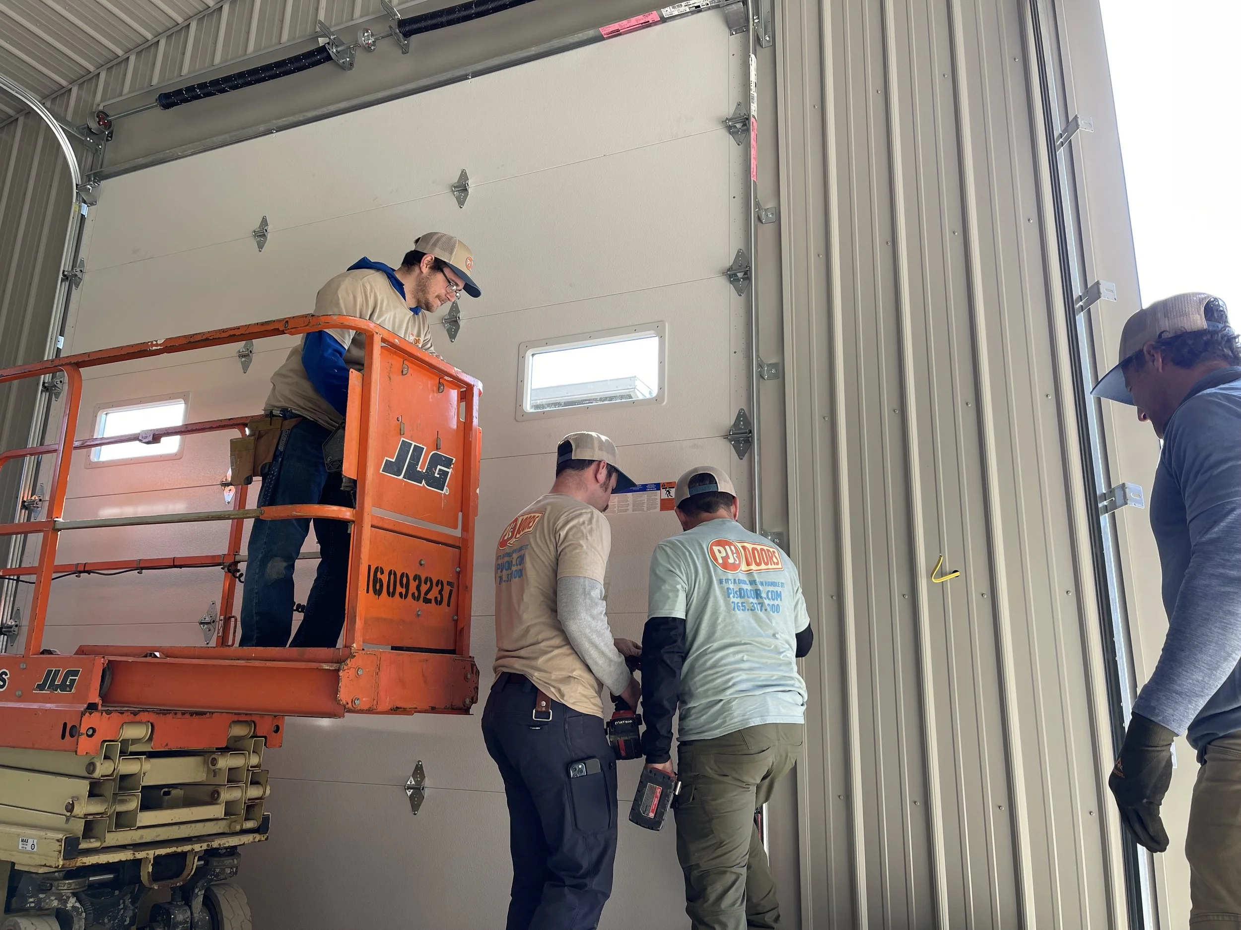 Two workers on a yellow scissor lift installing drywall on an overhead ceiling of a garage. One worker is wearing a black helmet, and the other is wearing glasses and a blue jacket. The garage door is open with a view of a parking lot and neighboring buildings.