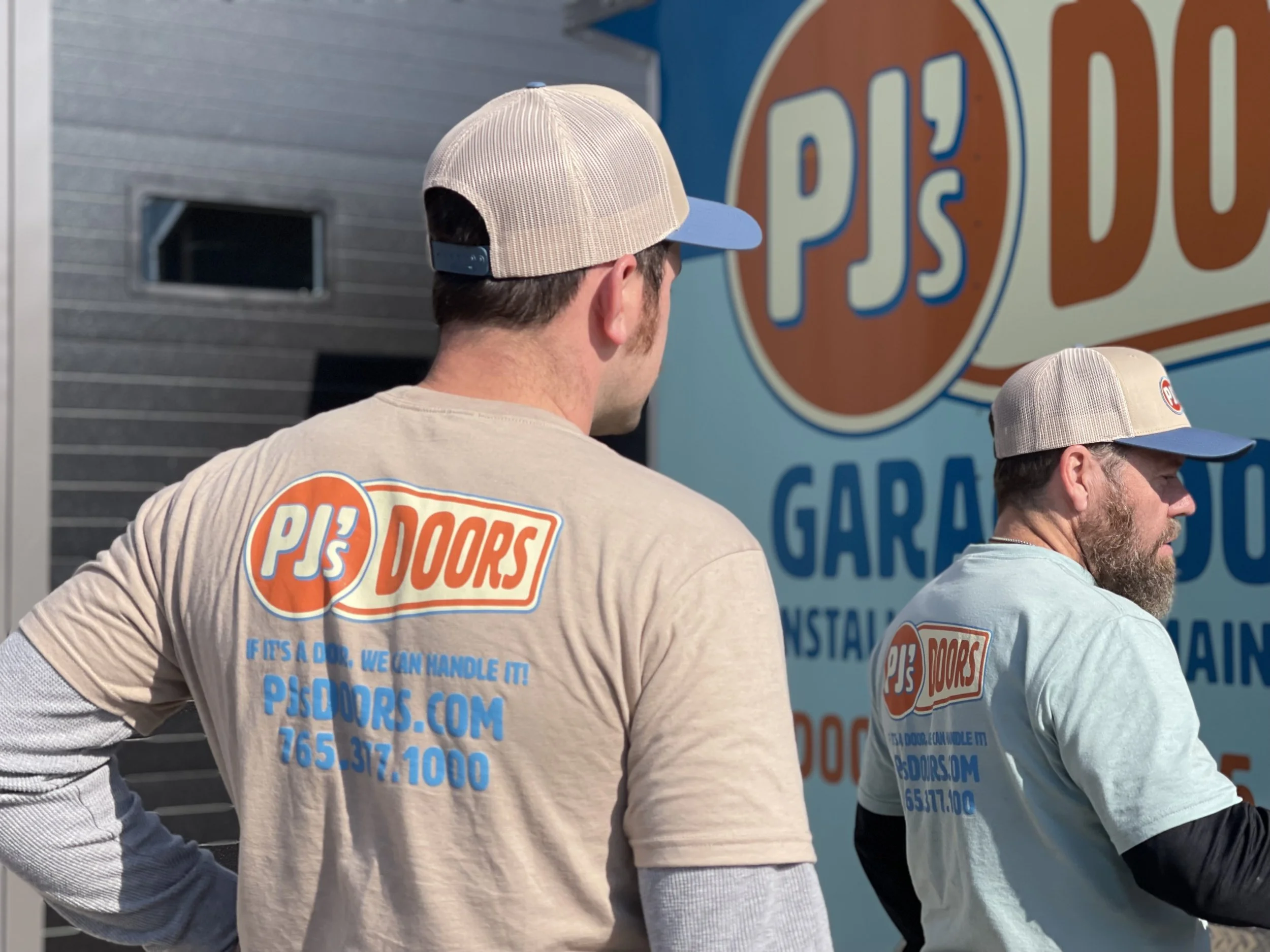 Two workers on a yellow scissor lift installing drywall on an overhead ceiling of a garage. One worker is wearing a black helmet, and the other is wearing glasses and a blue jacket. The garage door is open with a view of a parking lot and neighboring buildings.