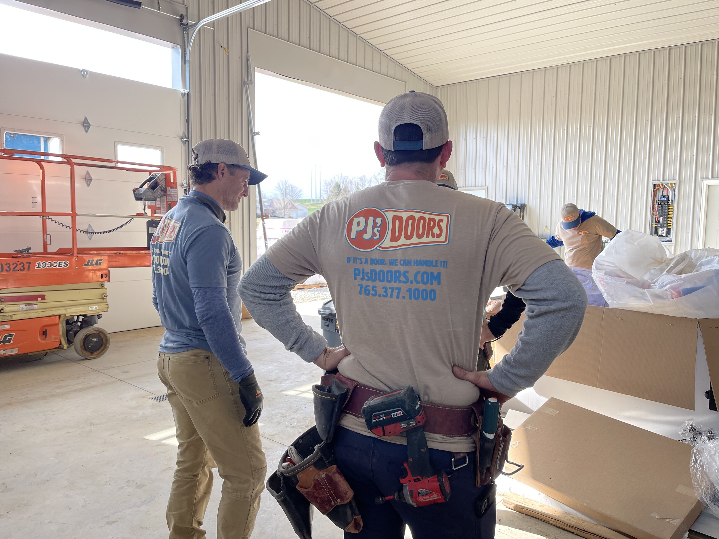 Two workers on a yellow scissor lift installing drywall on an overhead ceiling of a garage. One worker is wearing a black helmet, and the other is wearing glasses and a blue jacket. The garage door is open with a view of a parking lot and neighboring buildings.