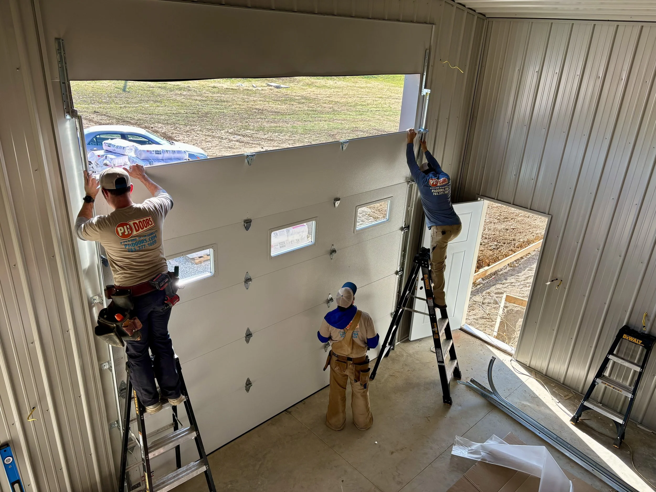 Three workers installing a large garage door inside an industrial building. One worker is on a ladder adjusting the upper part of the door, another is on a ladder securing the top edge, and the third is standing on the ground observing.
