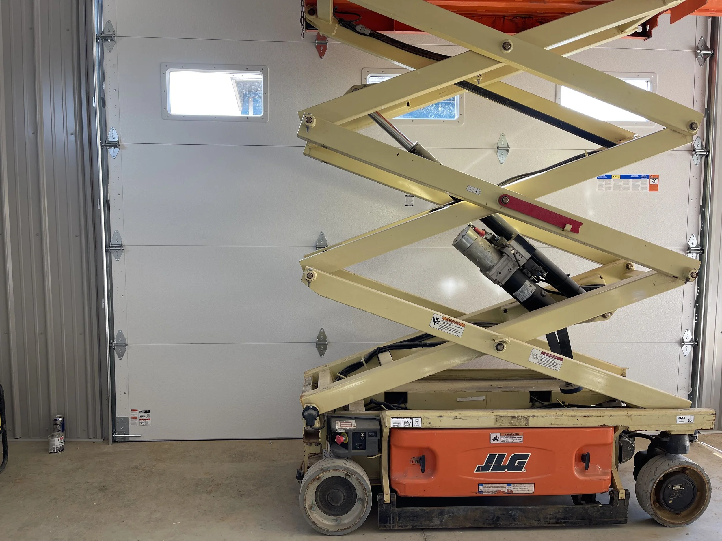 A yellow JLG scissor lift inside a garage, with a white garage door and small windows in the background.