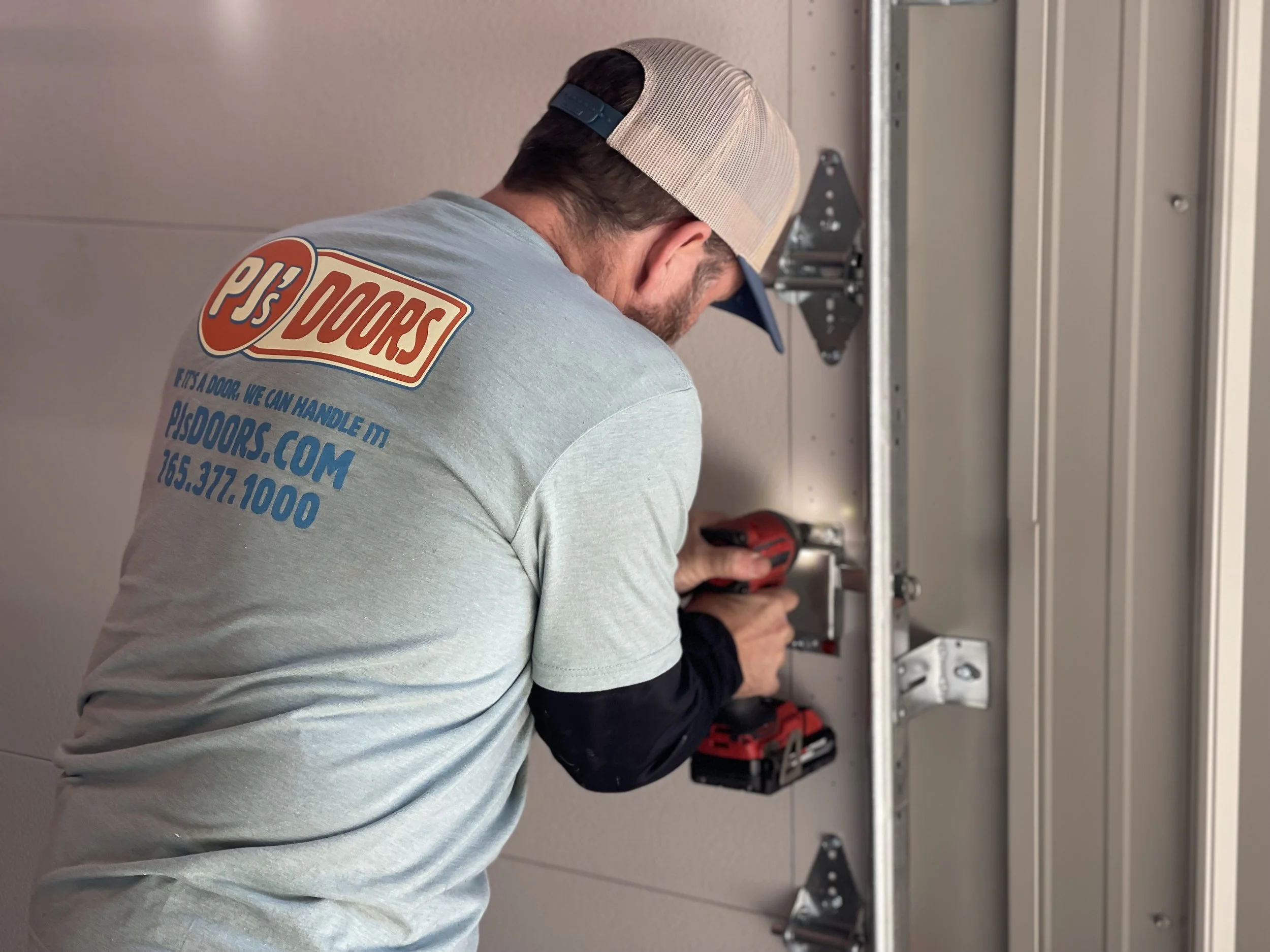 A man wearing a gray P's Doors t-shirt and a beige trucker hat is installing or repairing a garage door, using a power drill on the garage door hardware.