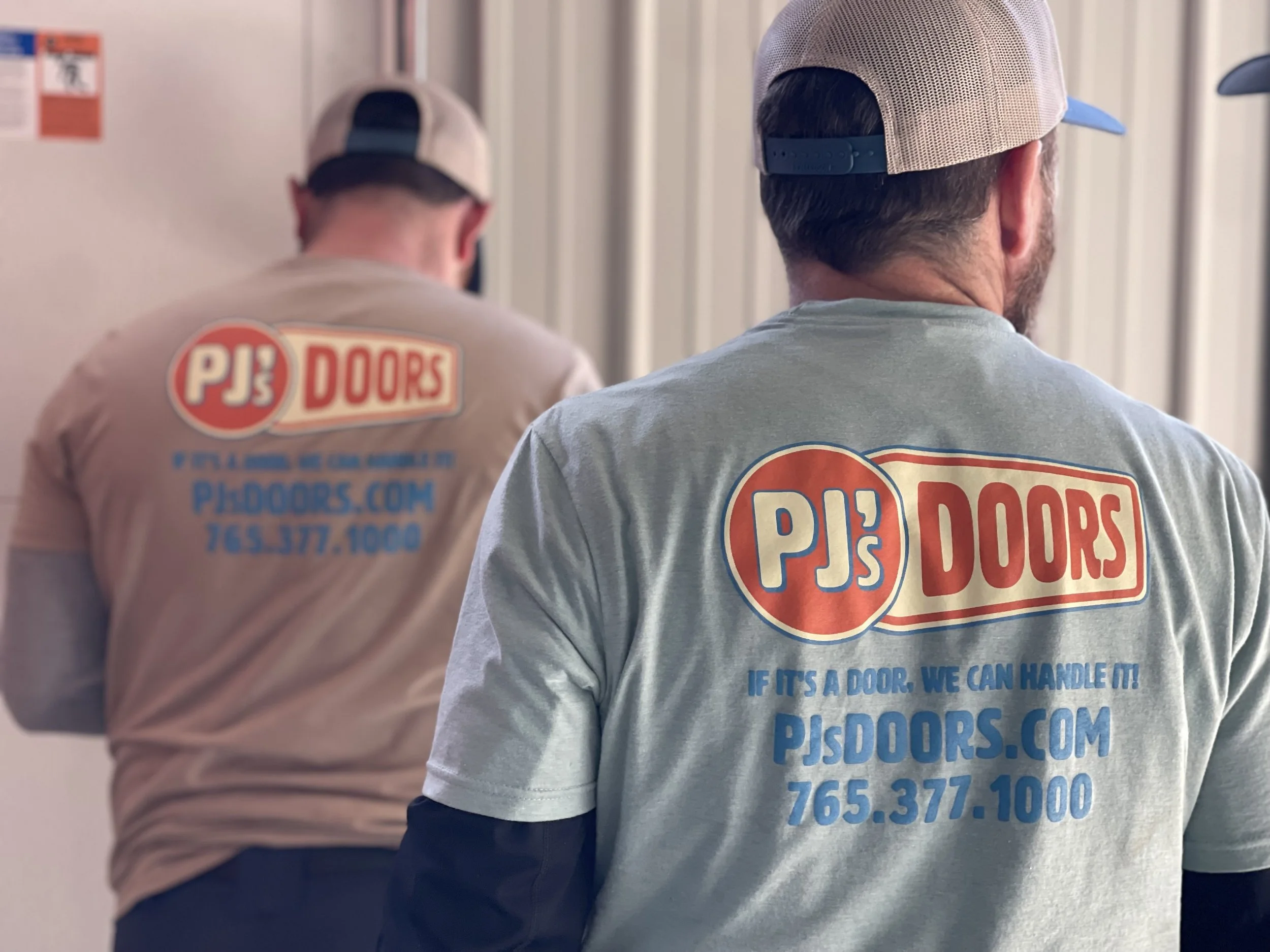 Two workers on a yellow scissor lift installing drywall on an overhead ceiling of a garage. One worker is wearing a black helmet, and the other is wearing glasses and a blue jacket. The garage door is open with a view of a parking lot and neighboring buildings.