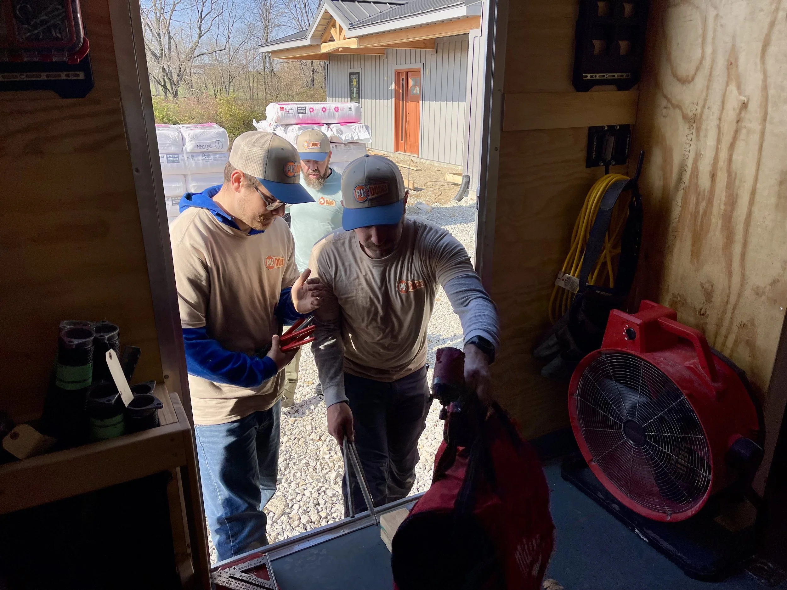 Two workers on a yellow scissor lift installing drywall on an overhead ceiling of a garage. One worker is wearing a black helmet, and the other is wearing glasses and a blue jacket. The garage door is open with a view of a parking lot and neighboring buildings.