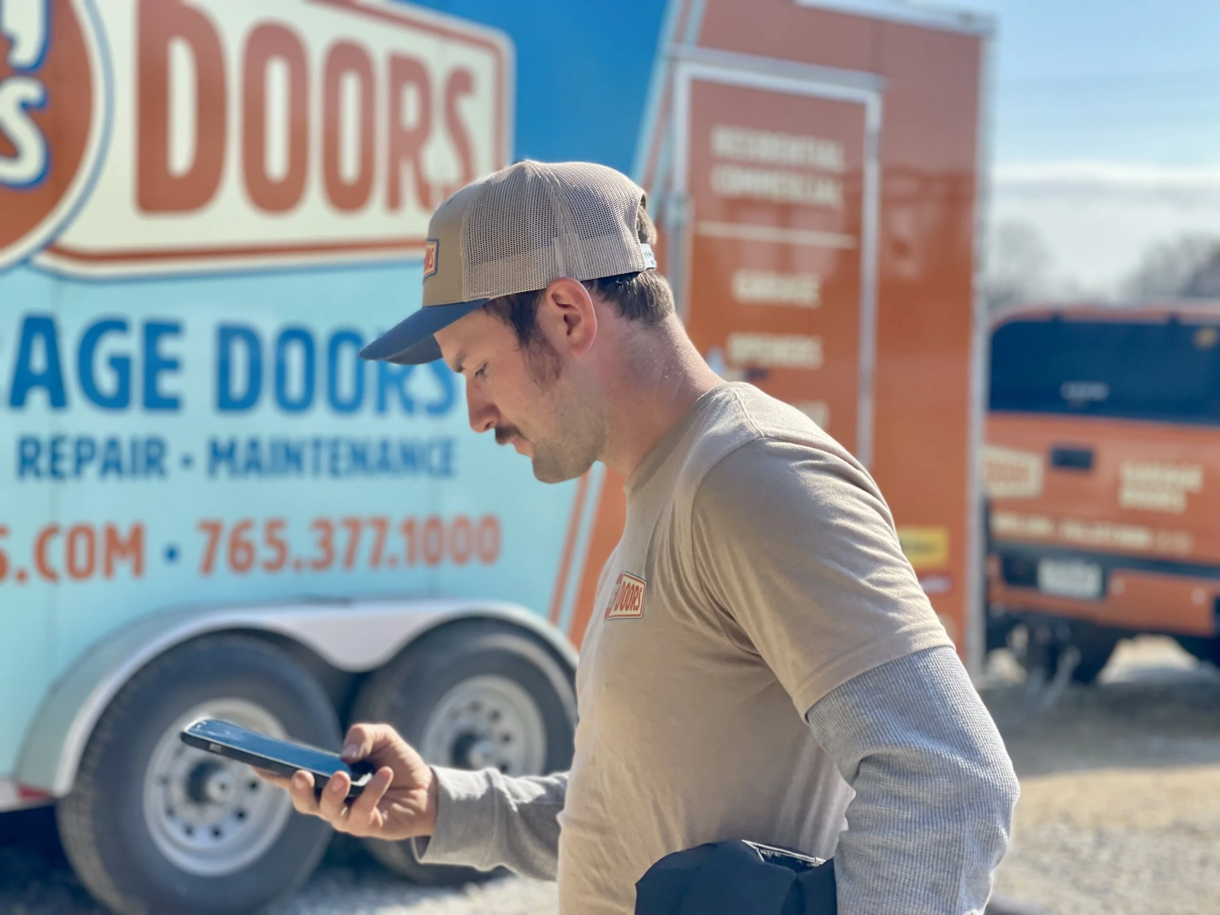 Two smiling men taking a selfie indoors, with a garage door in the background. One wears a black cap, and the other wears a gray cap with a 'New Horizons' logo.