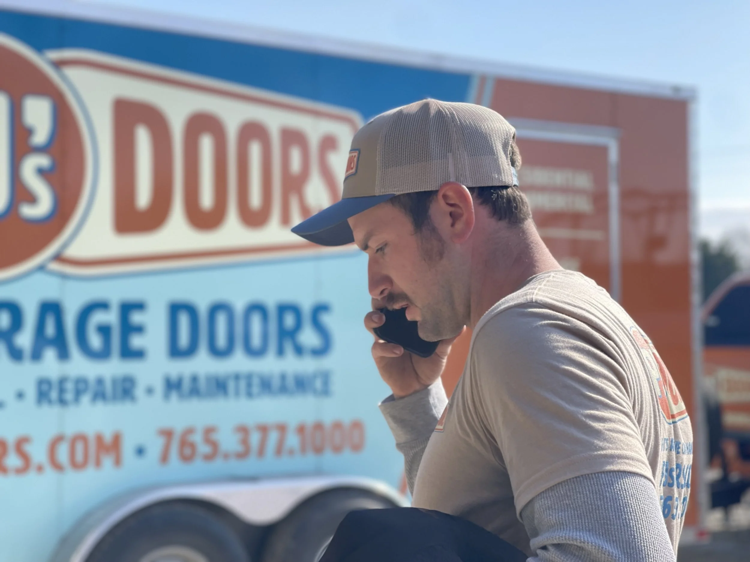 Two smiling men taking a selfie indoors, with a garage door in the background. One wears a black cap, and the other wears a gray cap with a 'New Horizons' logo.