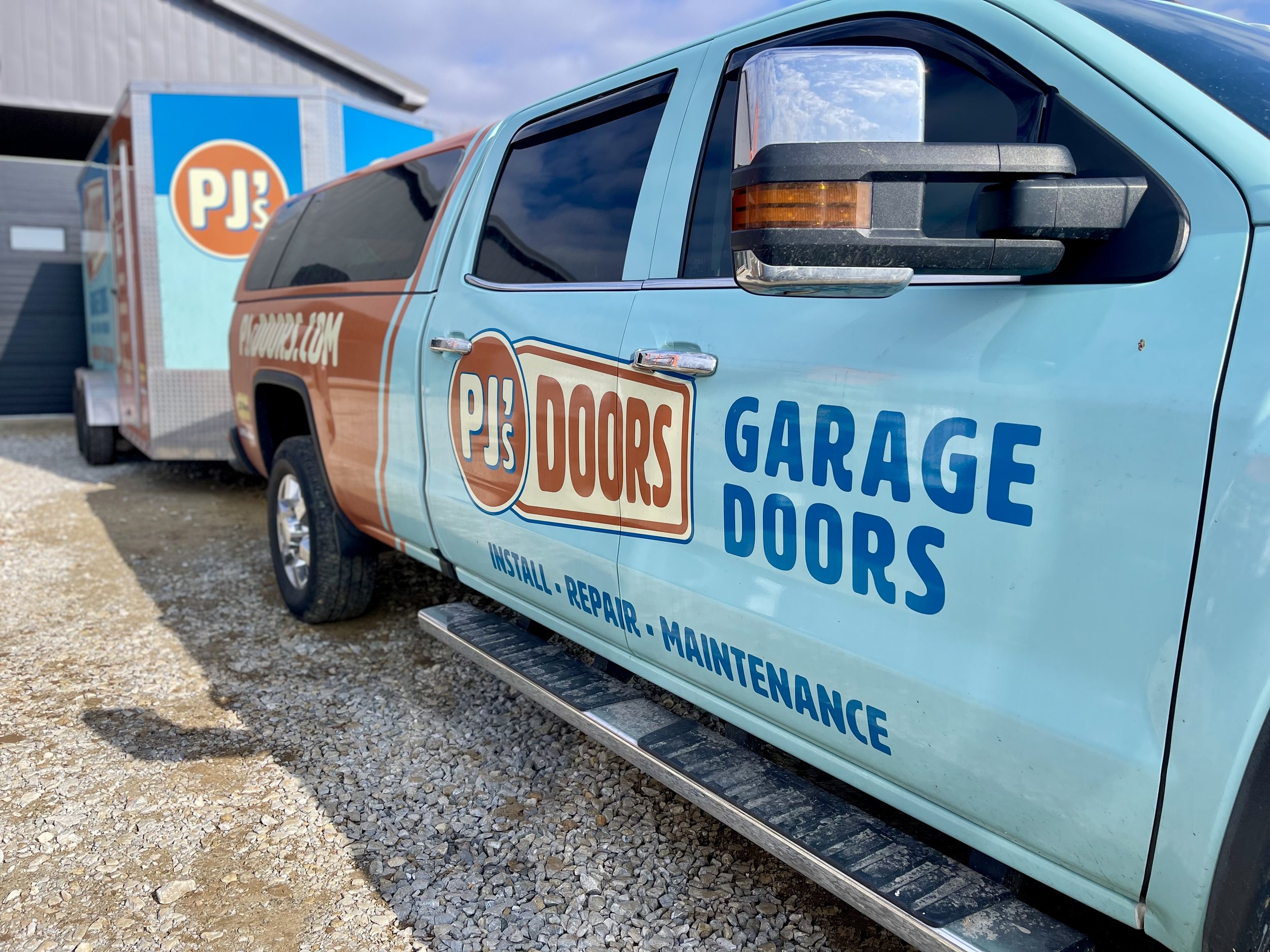 Two smiling men taking a selfie indoors, with a garage door in the background. One wears a black cap, and the other wears a gray cap with a 'New Horizons' logo.