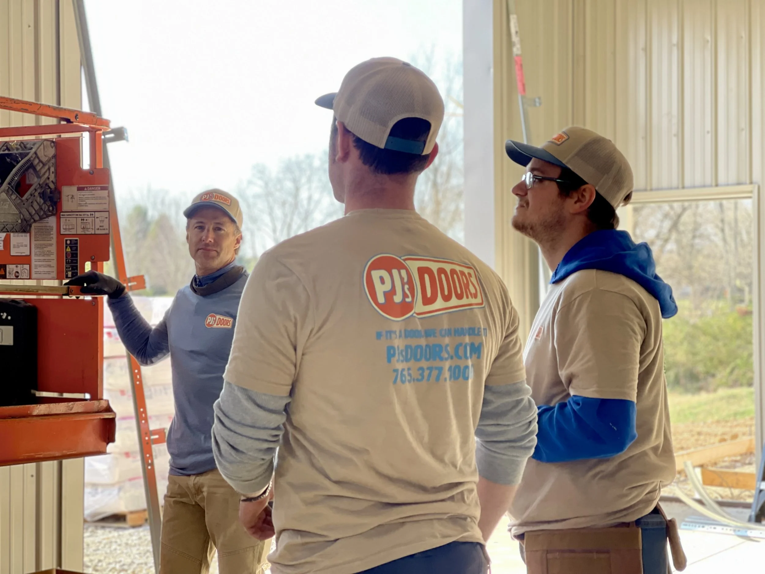 Three construction workers in a workshop, wearing hats and shirts with PJJ Doors logo, engaging in a discussion, with one worker gesturing near tools and equipment.