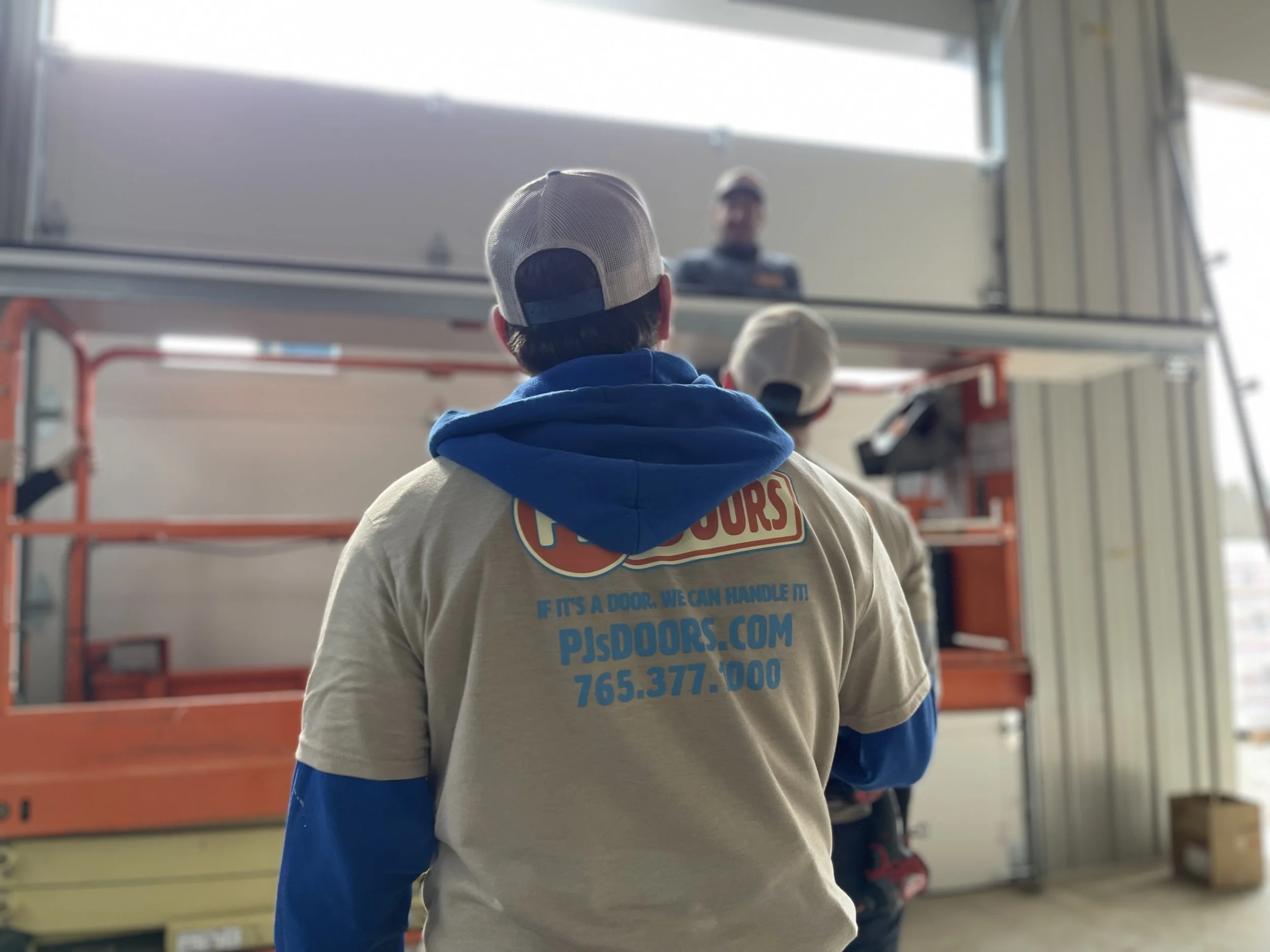 Two smiling men taking a selfie indoors, with a garage door in the background. One wears a black cap, and the other wears a gray cap with a 'New Horizons' logo.
