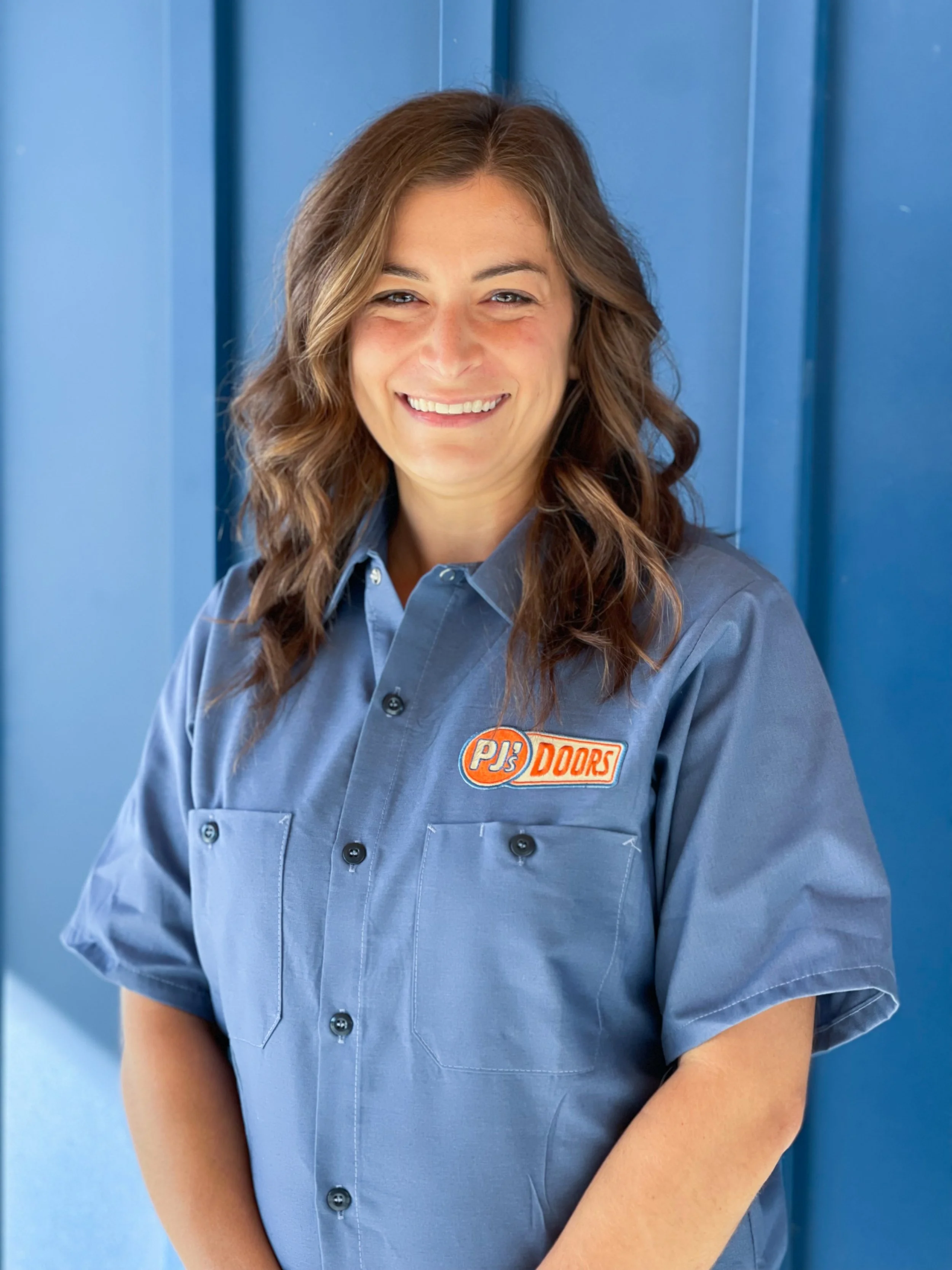 A woman with wavy brown hair smiling, wearing a beige T-shirt with a Pj's Doors logo, standing in front of a blue wall.