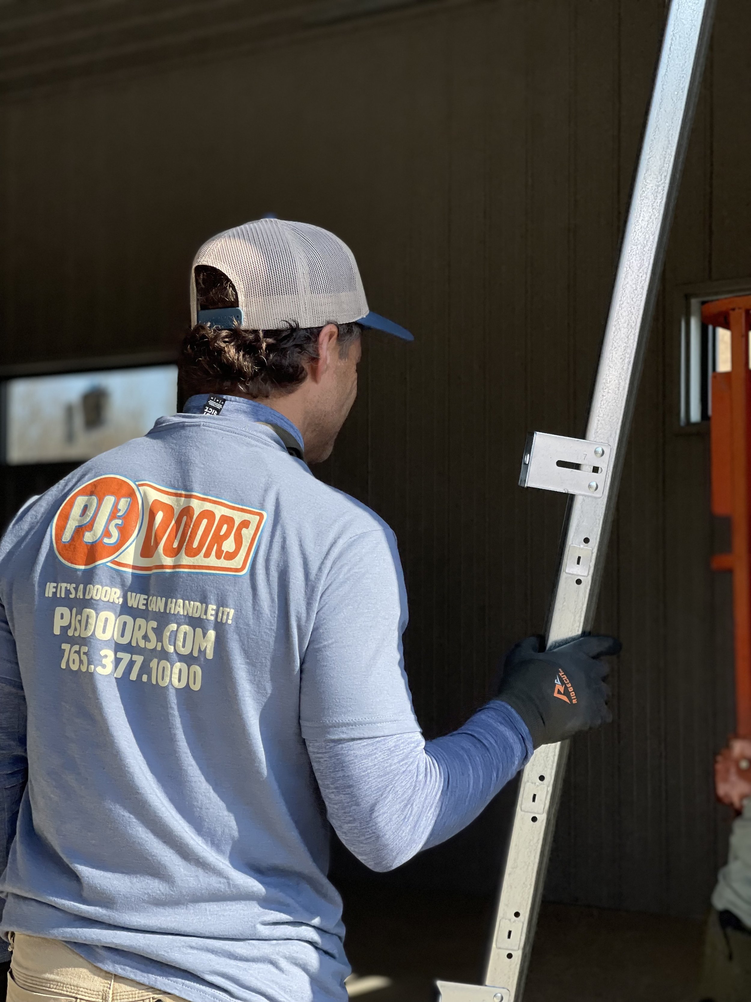 A man wearing a grey PJ's Doors shirt, a grey and blue baseball cap, and black gloves is holding a ladder.