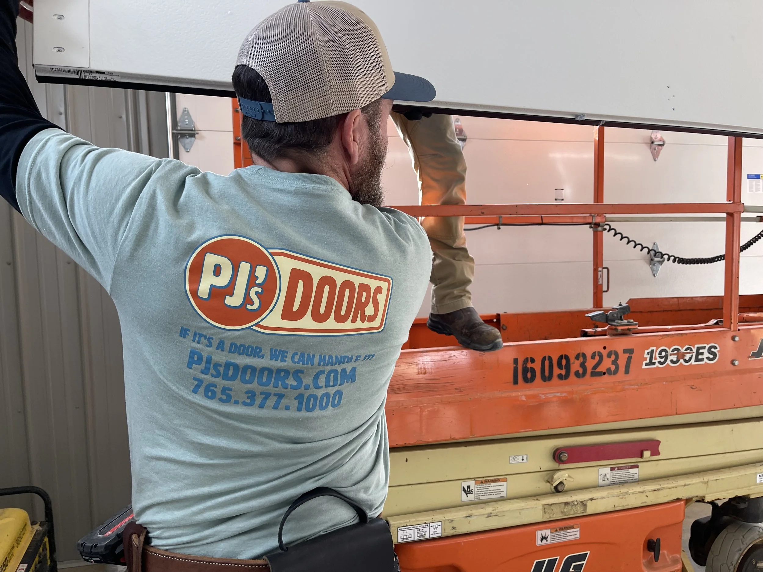 Two workers on a yellow scissor lift installing drywall on an overhead ceiling of a garage. One worker is wearing a black helmet, and the other is wearing glasses and a blue jacket. The garage door is open with a view of a parking lot and neighboring buildings.