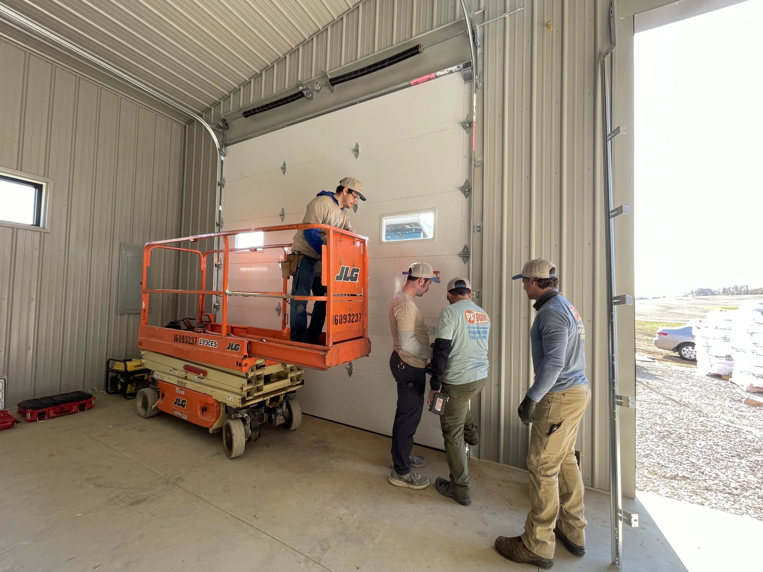 Four workers installing or repairing a large garage door inside a metal-paneled building using a scissor lift.