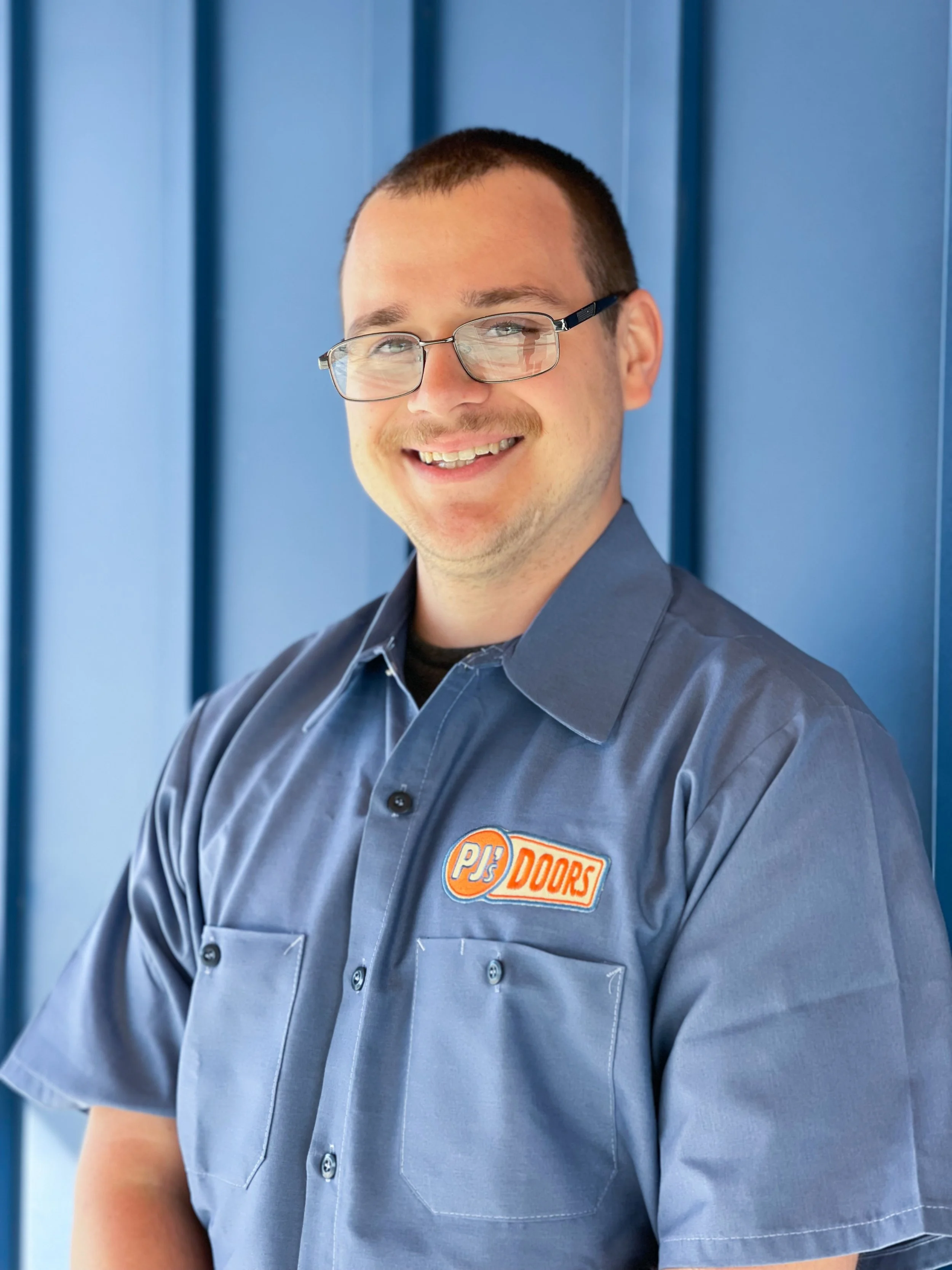 A smiling young man with glasses and a beige P.J.'s Doors t-shirt standing against a blue wall.