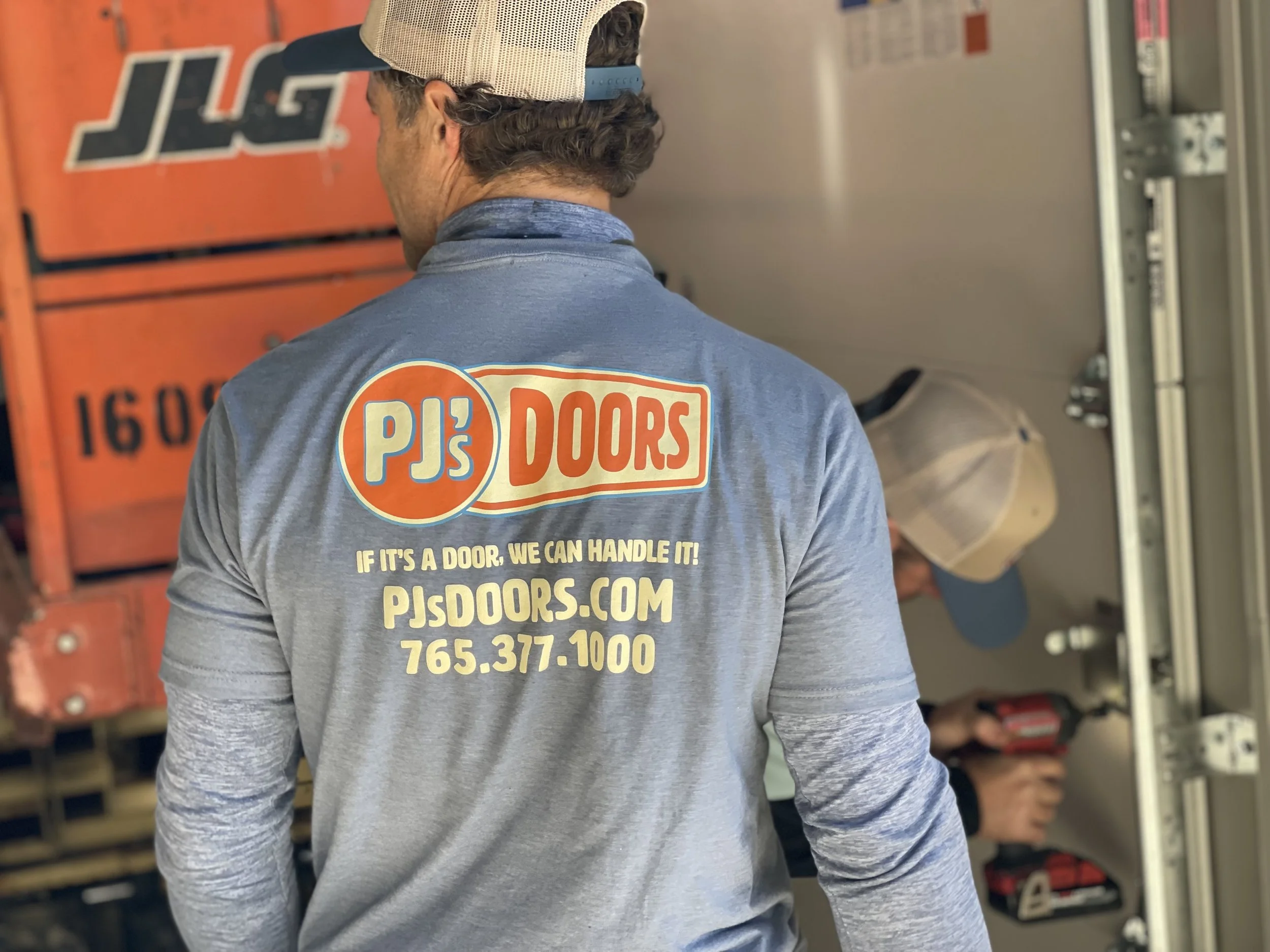 Two smiling men taking a selfie indoors, with a garage door in the background. One wears a black cap, and the other wears a gray cap with a 'New Horizons' logo.
