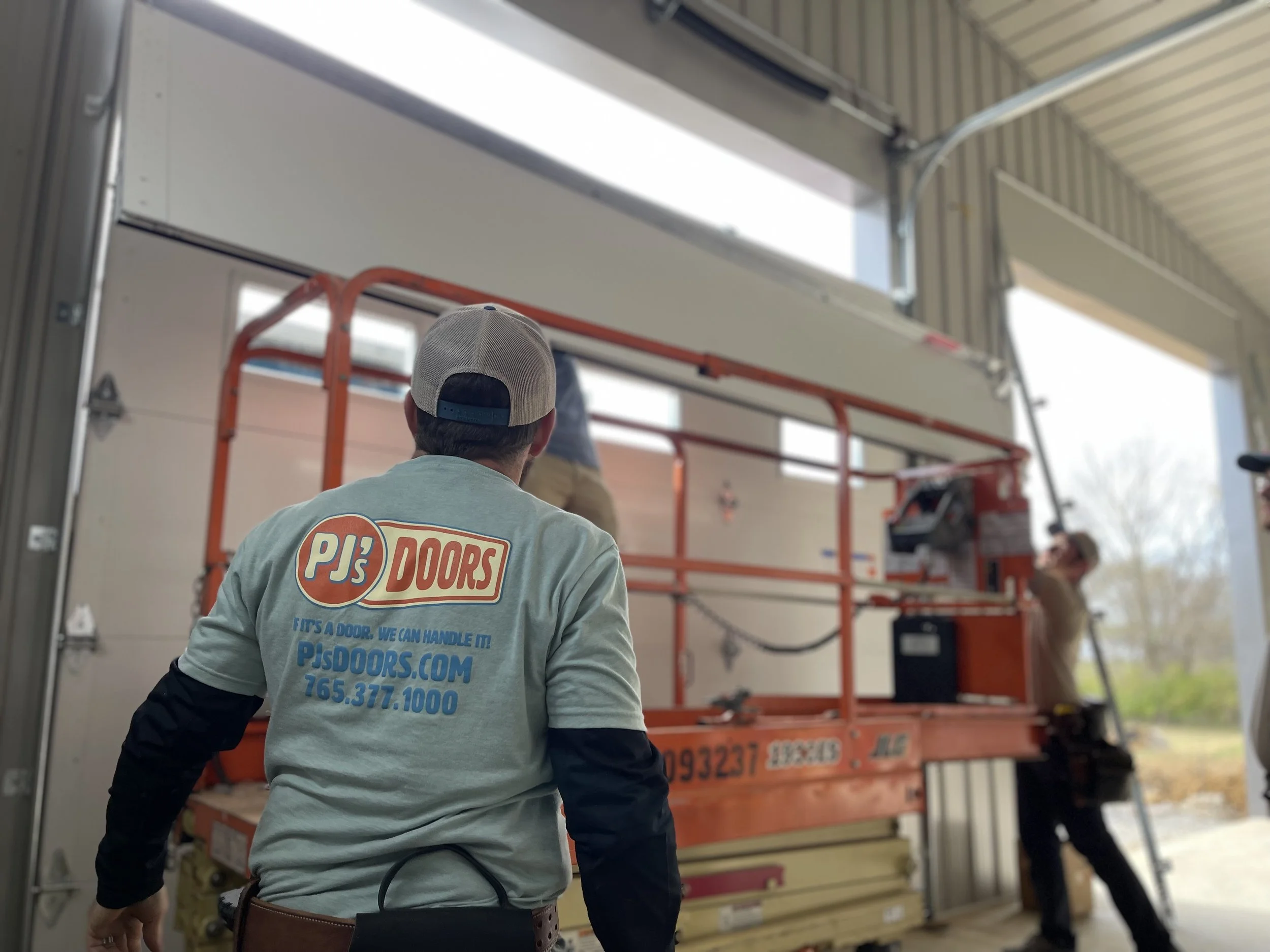 Workers installing a garage door inside a building, with one worker facing away wearing a PJ's Doors shirt and cap, another worker standing on a scissor lift, and an open garage door revealing outside scenery.