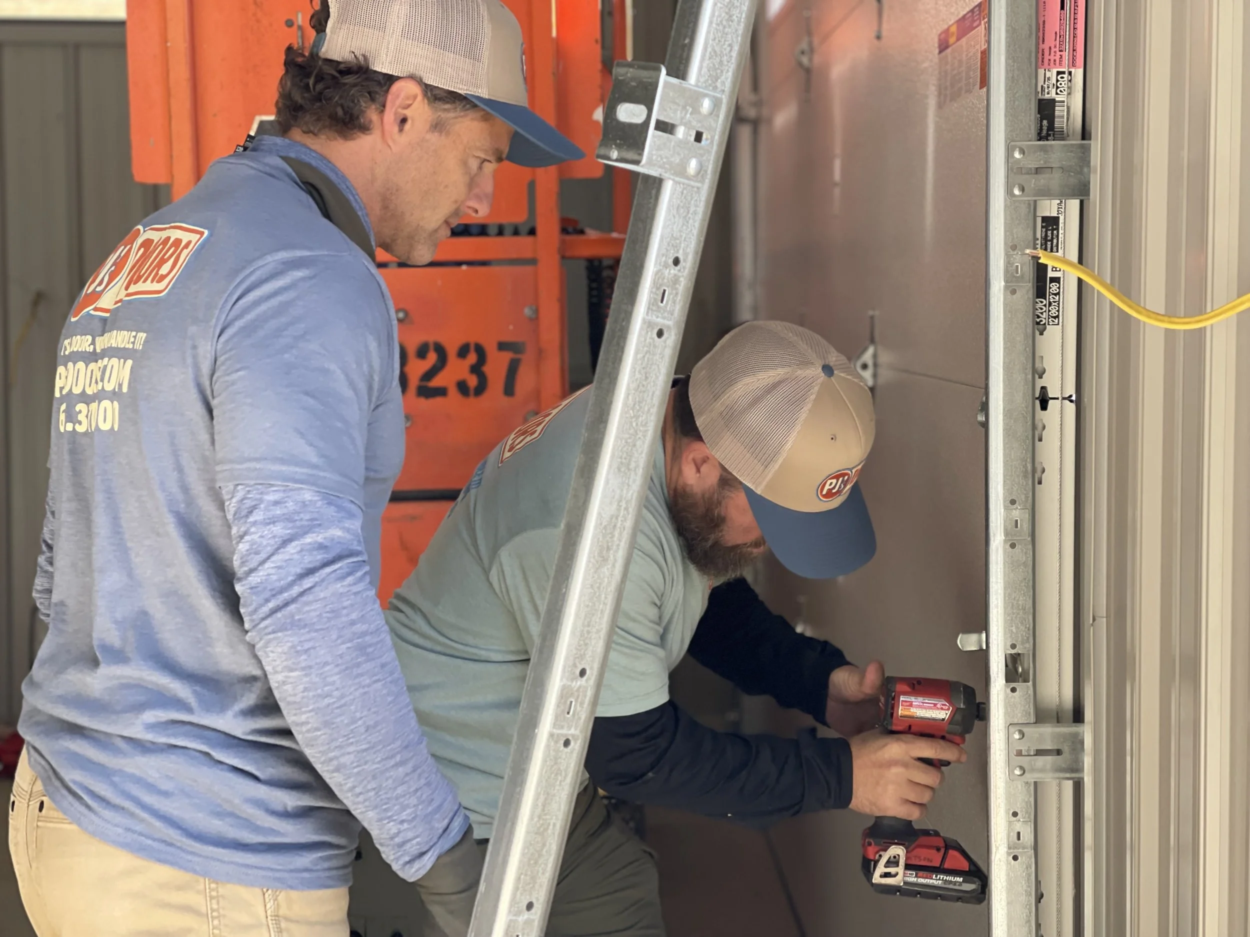 Two workers on a yellow scissor lift installing drywall on an overhead ceiling of a garage. One worker is wearing a black helmet, and the other is wearing glasses and a blue jacket. The garage door is open with a view of a parking lot and neighboring buildings.