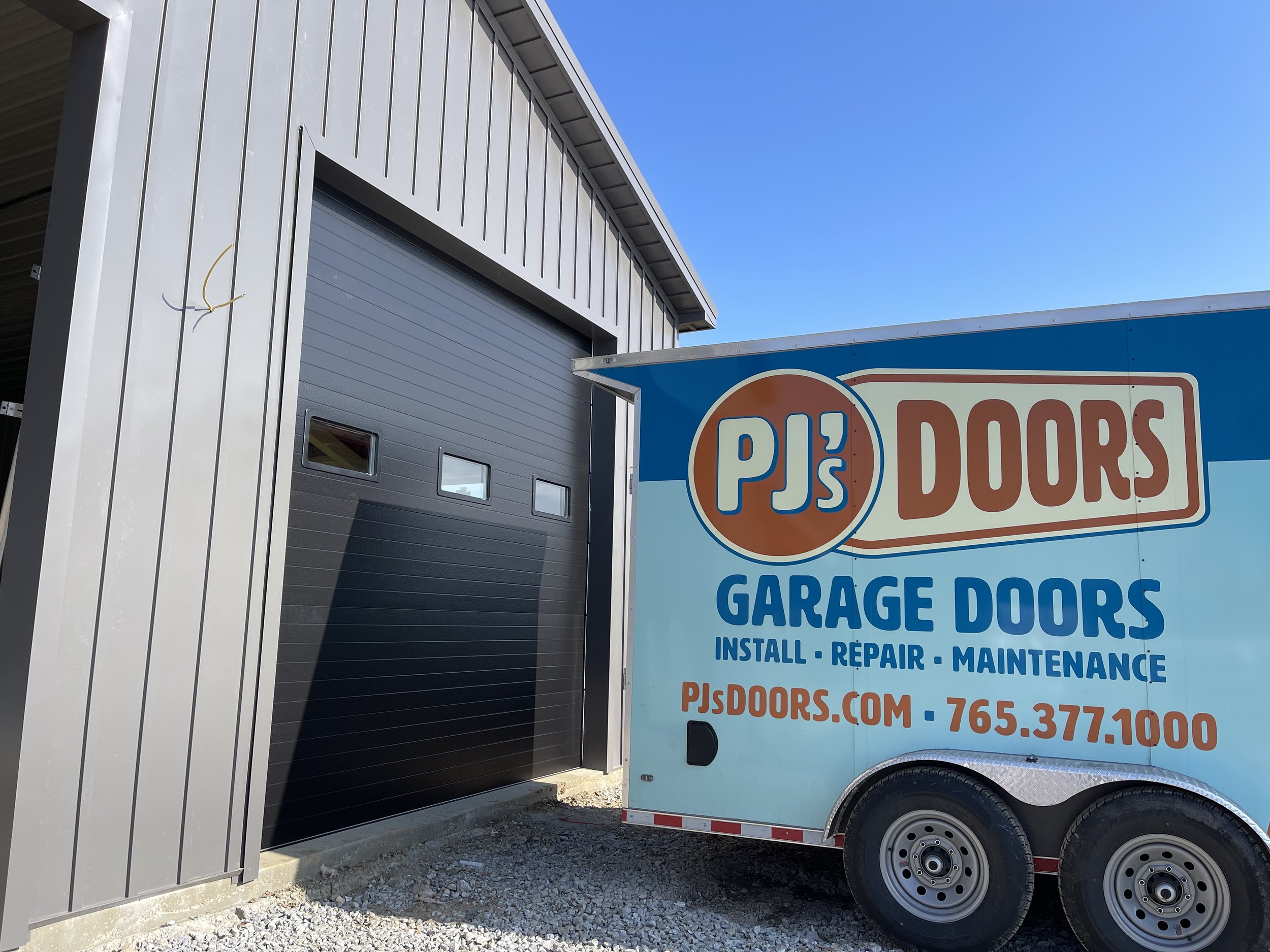 A grey garage door installed in a metal building, with a black mobile trailer nearby advertising PJ's Garage Doors, offering installation, repair, and maintenance services with a website and phone number.