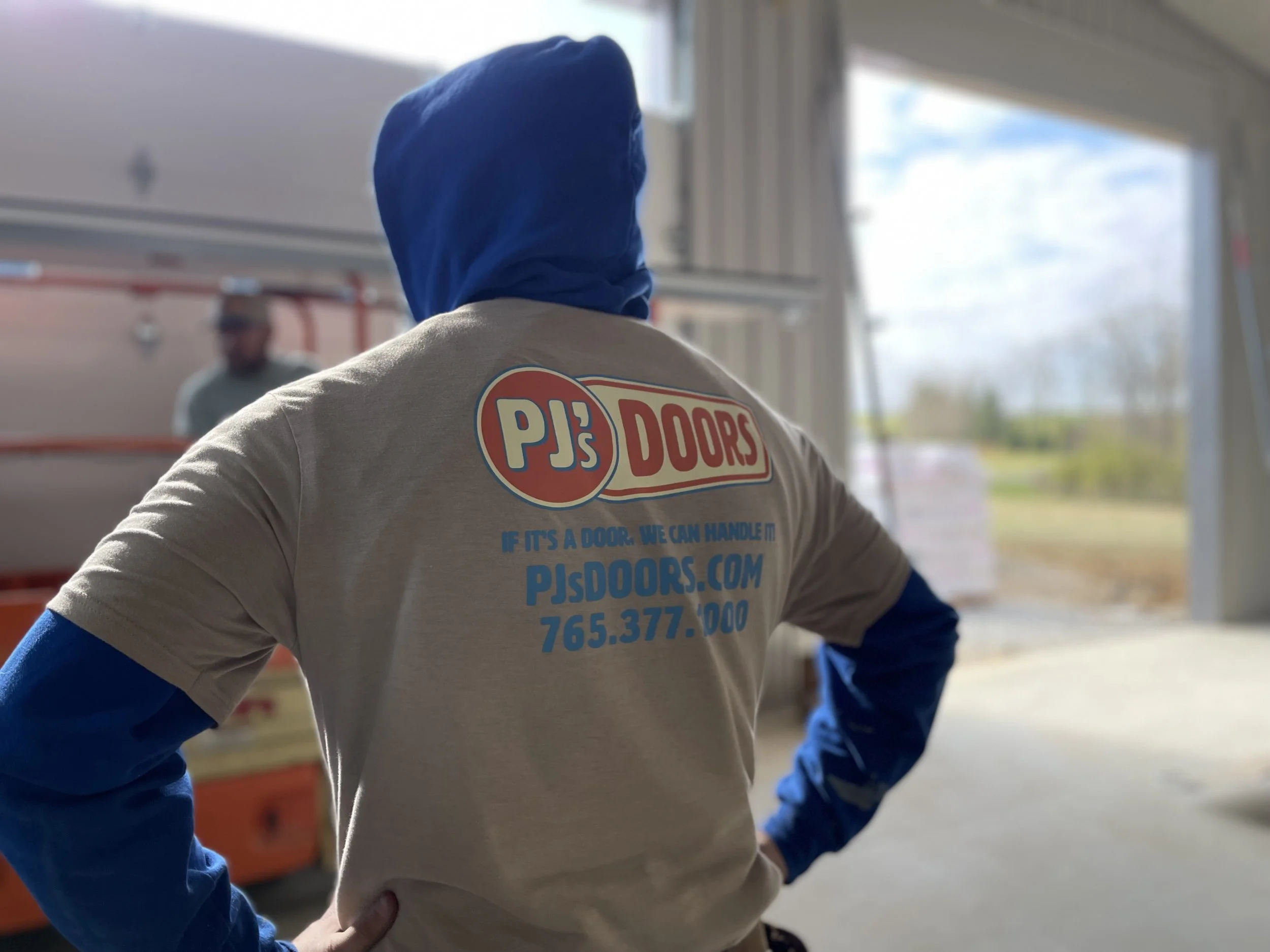 Person wearing a PJ's Doors t-shirt with a blue hood and blue sleeves, standing inside a garage or warehouse, with another person blurred in the background near an open door.