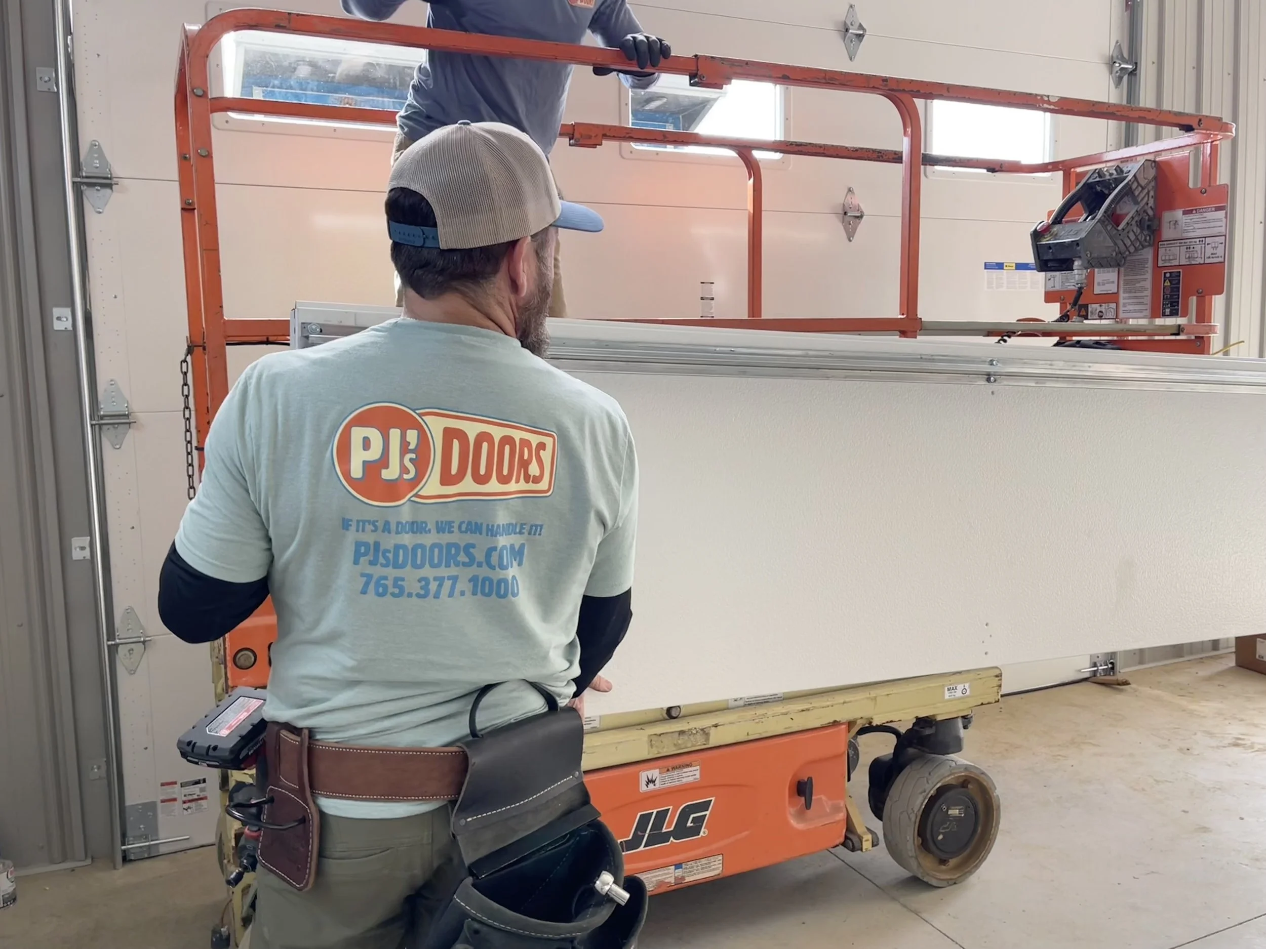 A worker wearing a PJ's Doors logo T-shirt and a baseball cap is operating an orange scissor lift while installing a white door inside a garage.