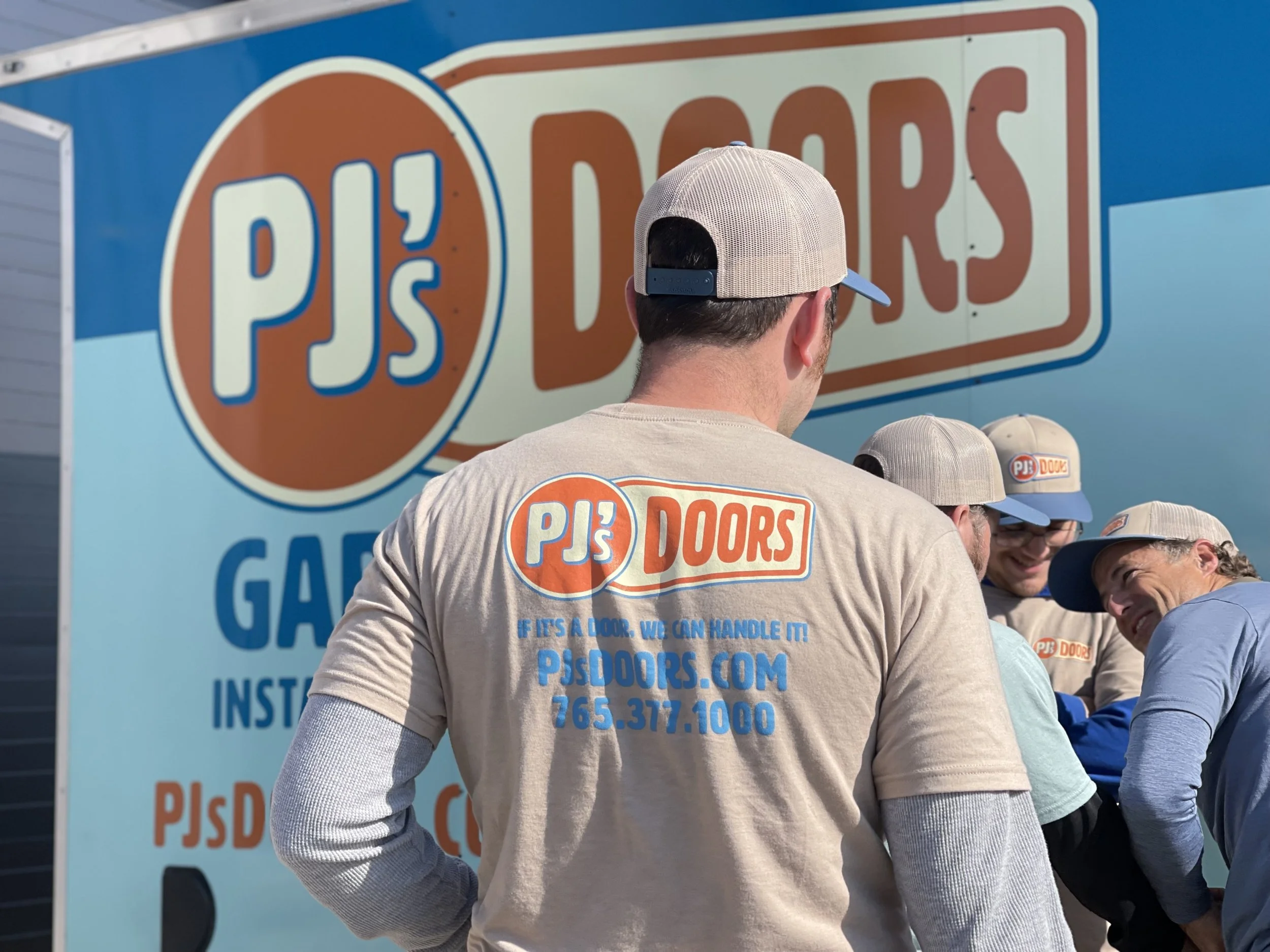 A group of men wearing matching beige PJ's Doors T-shirts and beige baseball caps stand in front of a large PJ's Doors sign. The sign features a blue and orange logo and text promoting their garage door services.