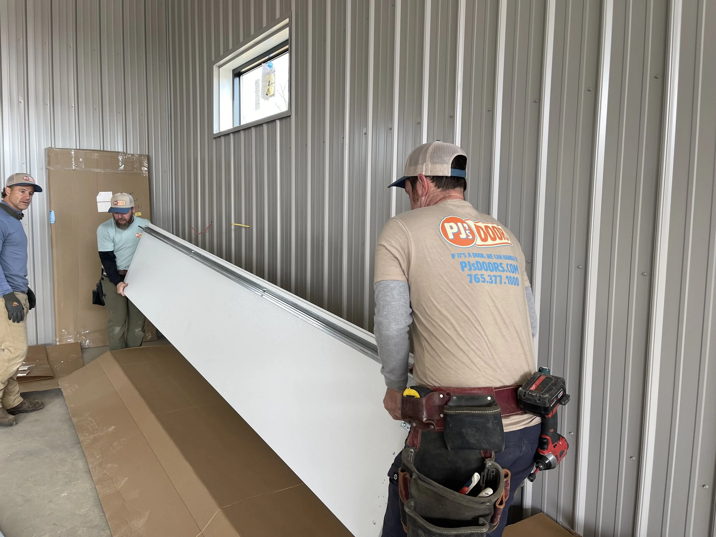 Three workers in a metal-paneled room installing a white door. One worker is holding the door, while the others stand nearby observing. The room has a small high window, and there are cardboard boxes and packaging materials on the floor.