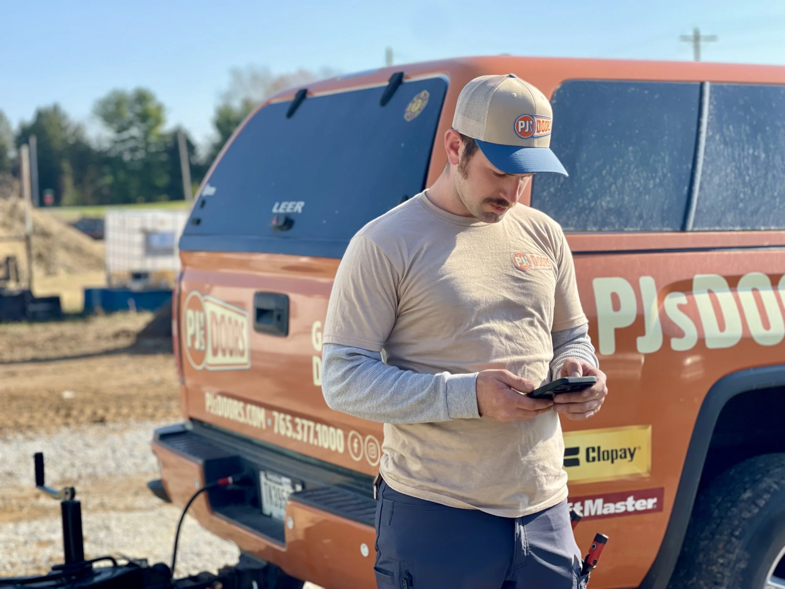 Two smiling men taking a selfie indoors, with a garage door in the background. One wears a black cap, and the other wears a gray cap with a 'New Horizons' logo.