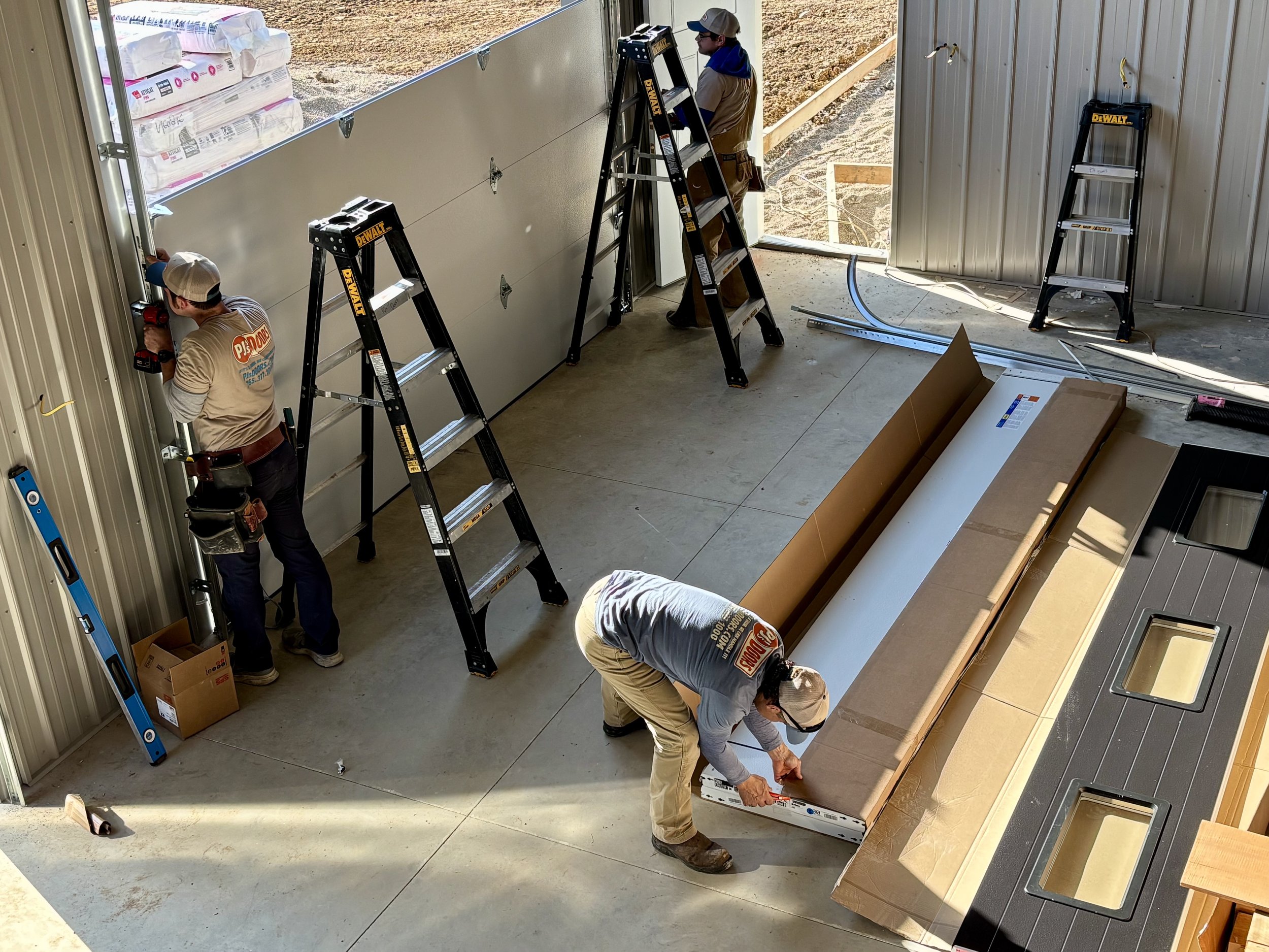 Two workers on a yellow scissor lift installing drywall on an overhead ceiling of a garage. One worker is wearing a black helmet, and the other is wearing glasses and a blue jacket. The garage door is open with a view of a parking lot and neighboring buildings.