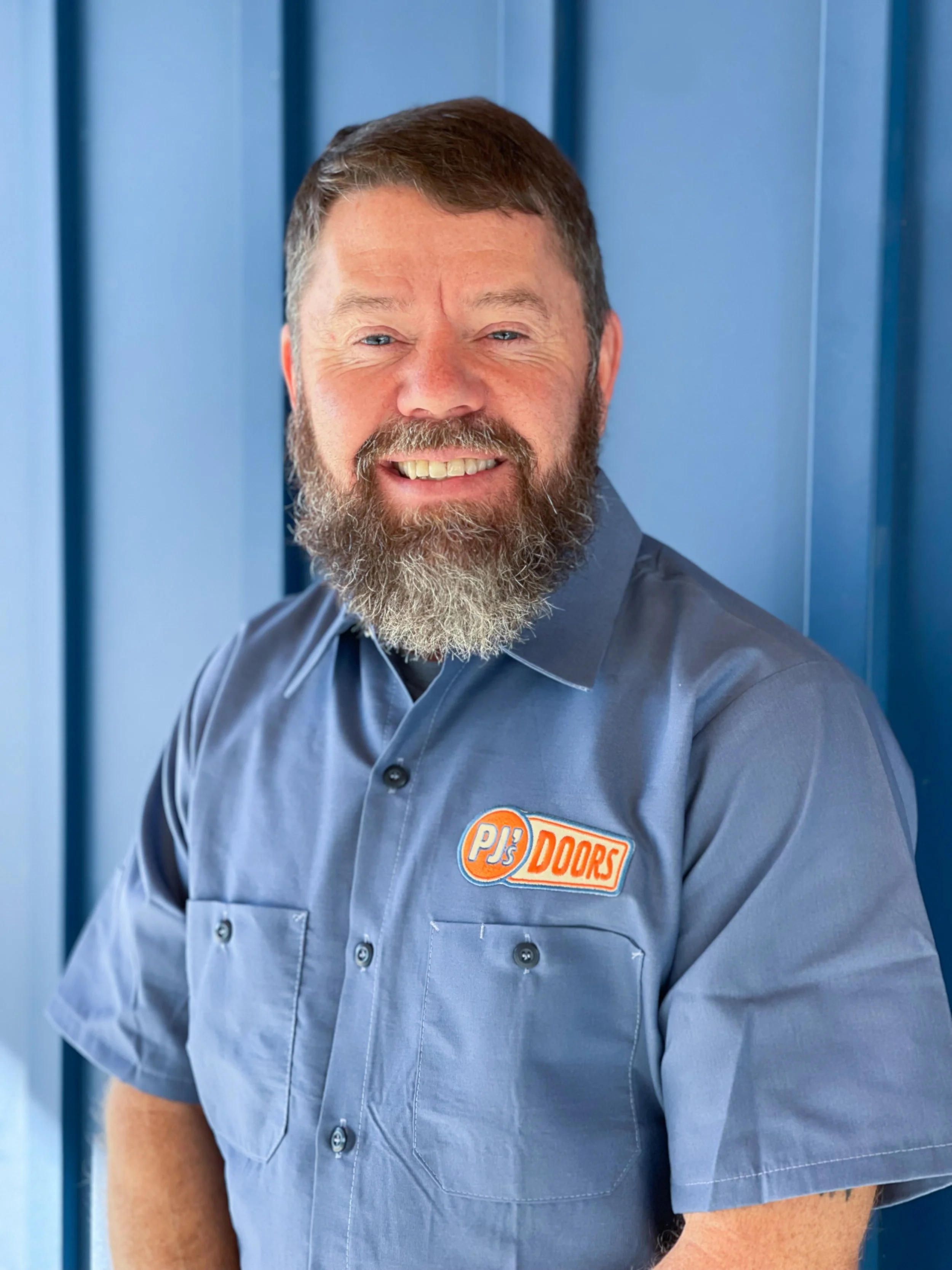 Man with a beard and mustache wearing a tan PJ's Doors T-shirt standing against a blue wall.
